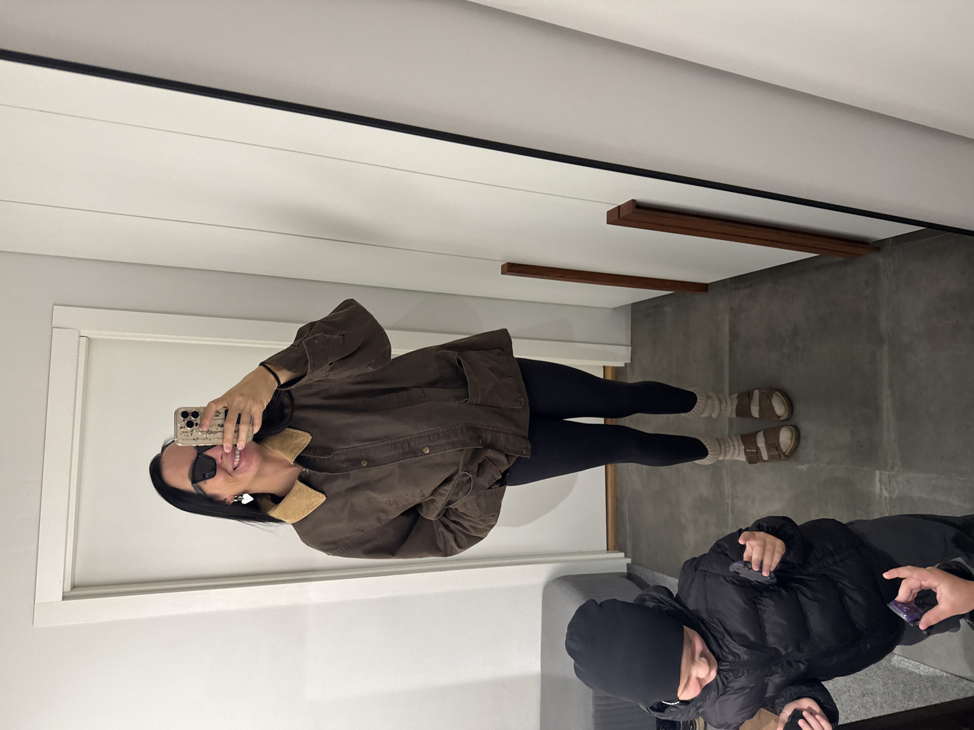 A woman stands in what appears to be a hallway or entryway, taking a mirror selfie. She is wearing a brown jacket with a corduroy collar, black leggings, and sandals. She has on black sunglasses and a heart-shaped earring. In the foreground, a young child wearing a black puffer jacket and a black beanie is visible. The child is holding small objects in their hands. The environment includes a white door on the left, a grey bench, and a tall white structure with vertical wooden accents on the right. The flooring is a dark grey tile. The image quality is somewhat low. There is no indication of the city of Chaikovsky, Russia.