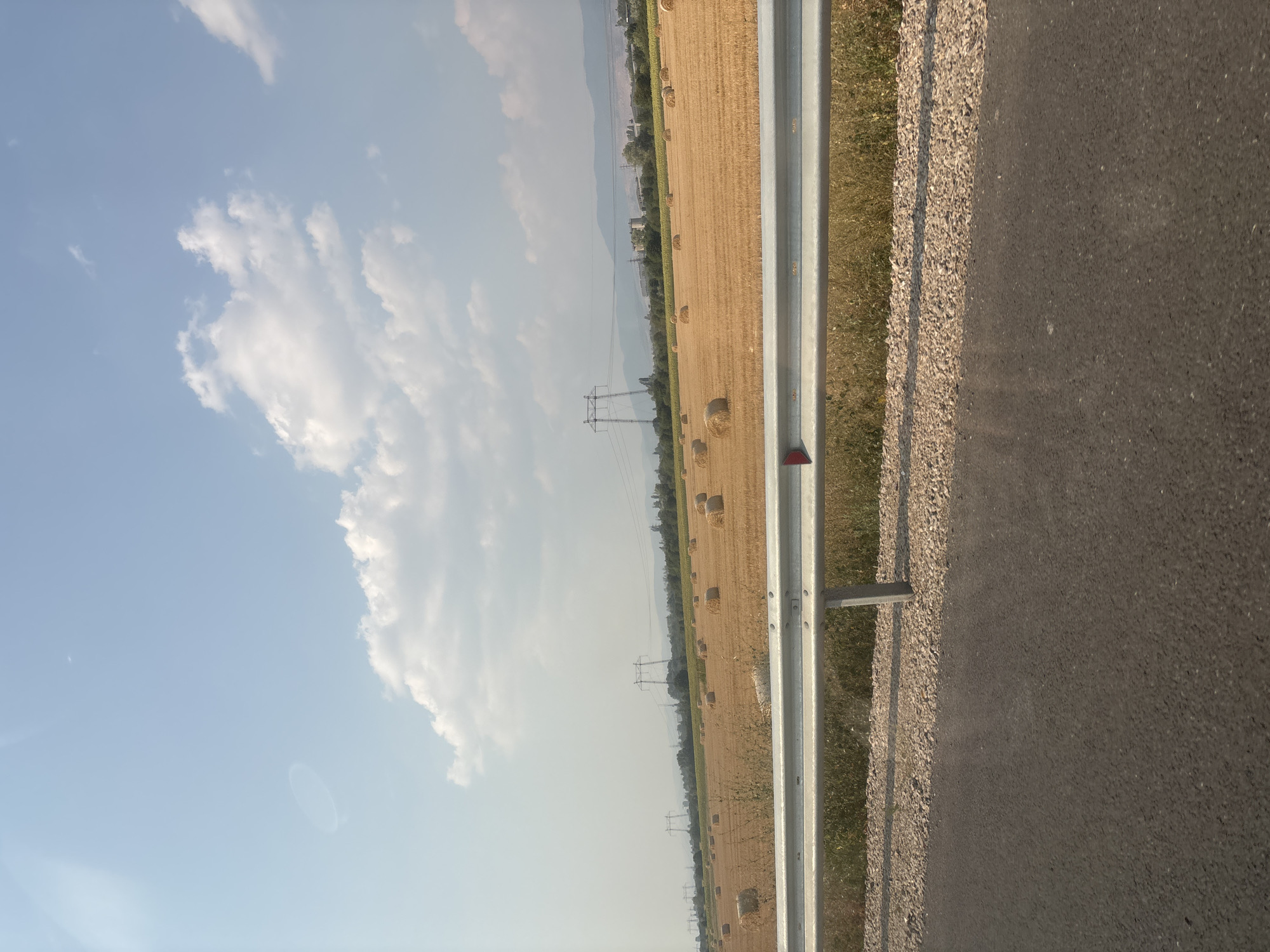 The image captures a rural landscape near Yana, Bulgaria, on a clear day. The foreground features an asphalt road and a metal guardrail, indicating the presence of a road. Beyond the guardrail, a vast field of harvested wheat is visible, dotted with numerous round hay bales, suggesting agricultural activity. In the middle ground, several tall power line towers stand prominently, with electrical wires stretching across the field, hinting at the proximity of infrastructure. The background reveals a line of trees and distant hills or mountains, under a bright blue sky with scattered white clouds. The overall scene depicts a serene, agricultural setting with signs of both nature and human-made structures.
