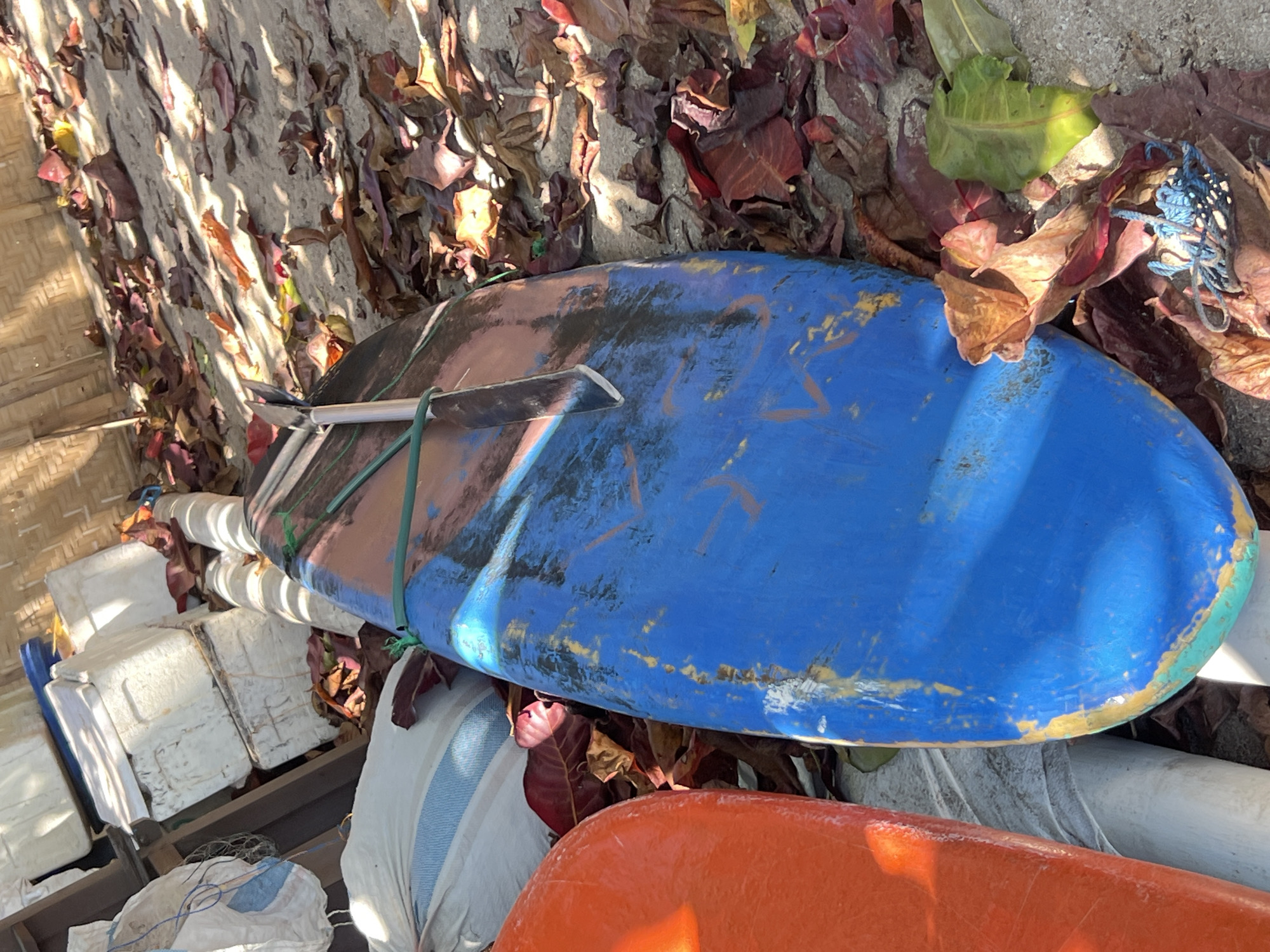 In Kuta, Indonesia, two surfboards lie on sandy ground amongst fallen leaves. One is bright orange and the other is blue with faded paint and some yellow markings. A silver fin is attached to the blue surfboard, secured with a green rope. Behind the surfboards, there are several white containers stacked, a large white sack with blue stripes, and other bags. A woven bamboo structure is visible in the background, suggesting an outdoor shelter or building. The ground is covered with a mixture of sand and dry, reddish-brown leaves, with some green leaves scattered among them. A tangled blue rope lies on the ground near the surfboards. The scene is lit by sunlight, casting shadows on the sand and leaves.