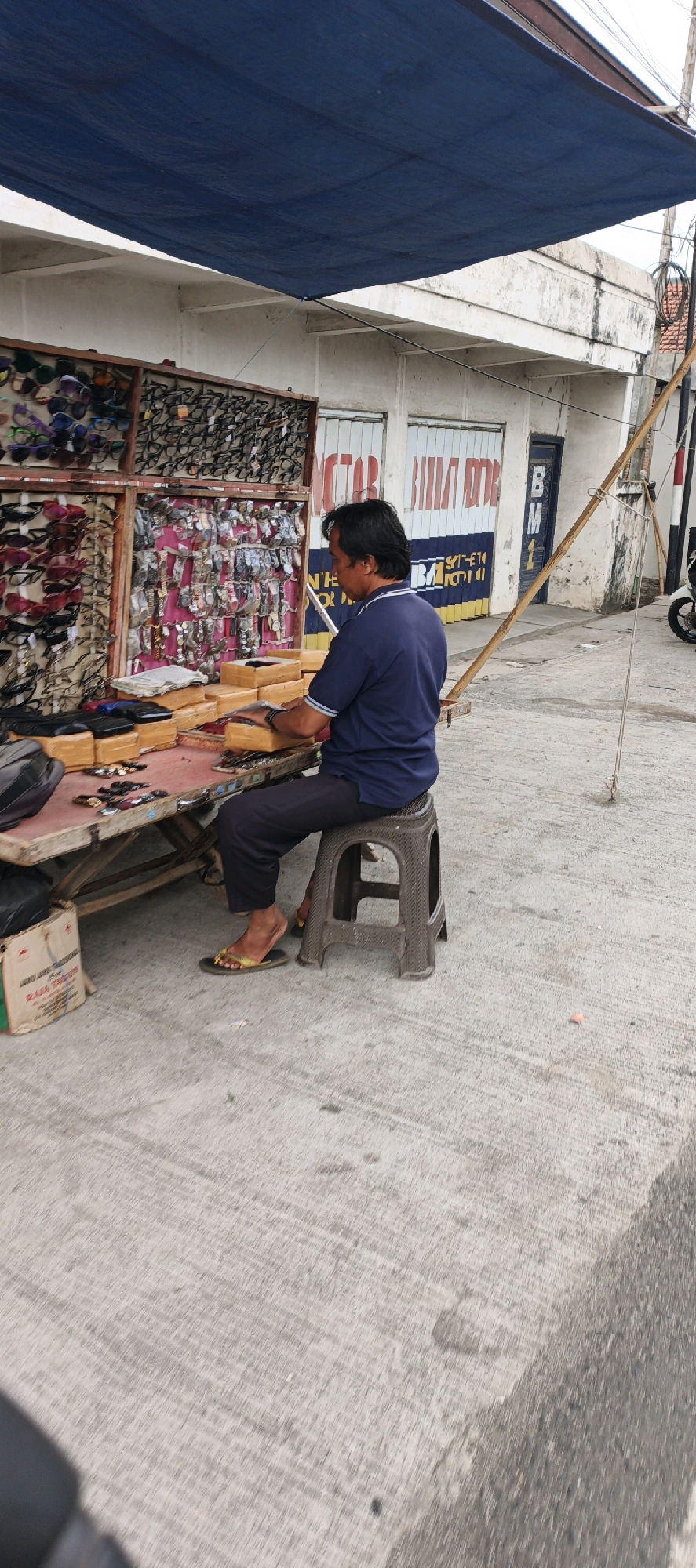 A man is sitting on a plastic stool at a roadside stall in Kasiyan, Indonesia, selling sunglasses and watches. He is wearing a blue t-shirt and dark pants. The stall is covered by a blue tarp, and displays are filled with numerous pairs of sunglasses and watches. The man is looking down at the merchandise on the table in front of him, which includes several boxes and individual watches. The ground is paved with concrete, and there is a building in the background with a sign that reads "VICTOR BILA DOOR".
