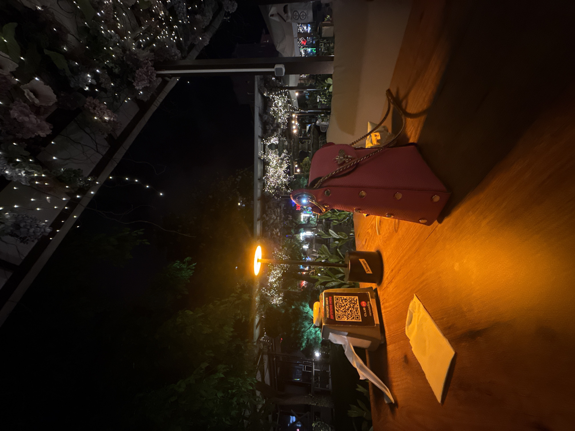 The image shows a table at an outdoor restaurant at night. On the table is a red purse, a QR code payment device with a tissue dispenser, and a crumpled white napkin. The table is illuminated by a warm light source, and in the background, there are decorative lights on trees and other foliage, suggesting an upscale dining atmosphere. The QR code on the payment device indicates that the restaurant is likely "Club 35" in Sofia, Bulgaria.