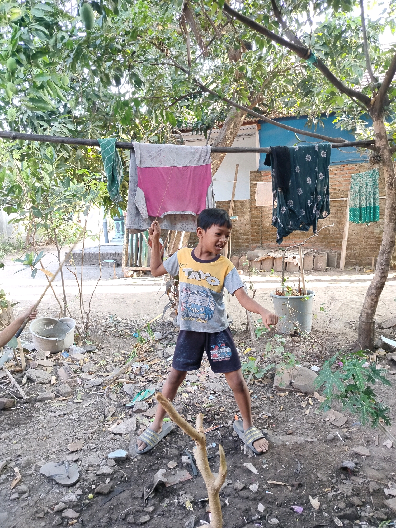 In Beloh, Indonesia, a young boy stands in a yard filled with debris and foliage. He is wearing a gray and yellow t-shirt with a graphic print and dark shorts. The boy is holding a string with both hands, appearing to be flying a kite or controlling a puppet. His mouth is open, and he looks to be engaged in an activity or shouting. In the background, laundry hangs on a clothesline strung between trees. The ground is uneven and covered with dirt, rocks, and scattered trash. A plastic bucket sits nearby. Several trees with leaves and branches are visible, some bearing fruit. Brick structures and a blue wall are partially visible behind the trees. The scene suggests an outdoor, informal setting, likely a residential backyard.