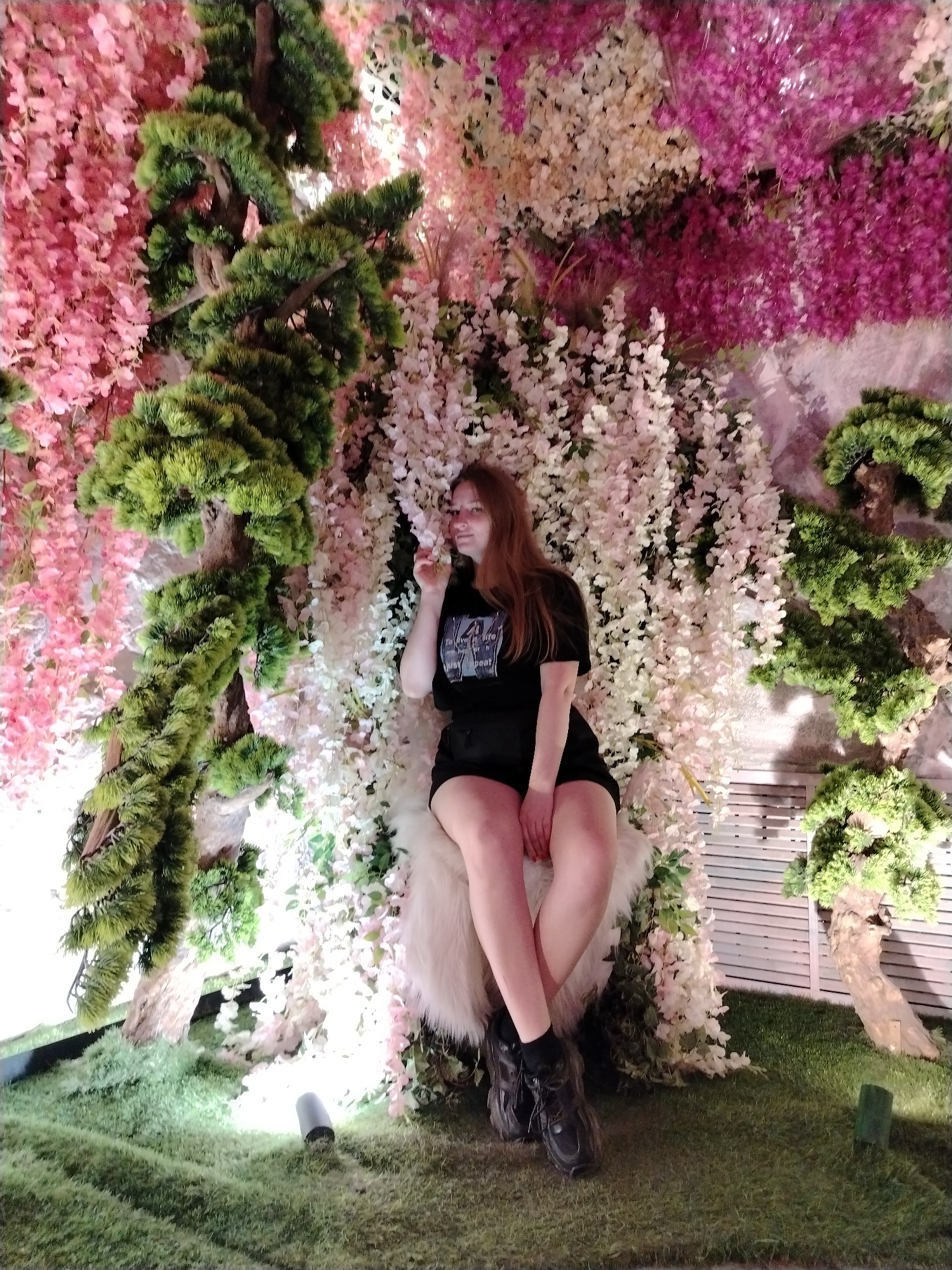 A young woman sits on a furry stool in an artificial floral display in Kazan, Russia. She is wearing a black t-shirt and shorts with black socks and dark sneakers. She is positioned in front of a wall covered in white and pink flowers, with a faux bonsai tree to her right and left. The ground is covered in artificial grass. A light fixture is visible on the ground near the tree to her left. The wall behind the floral display appears to be grey or white.