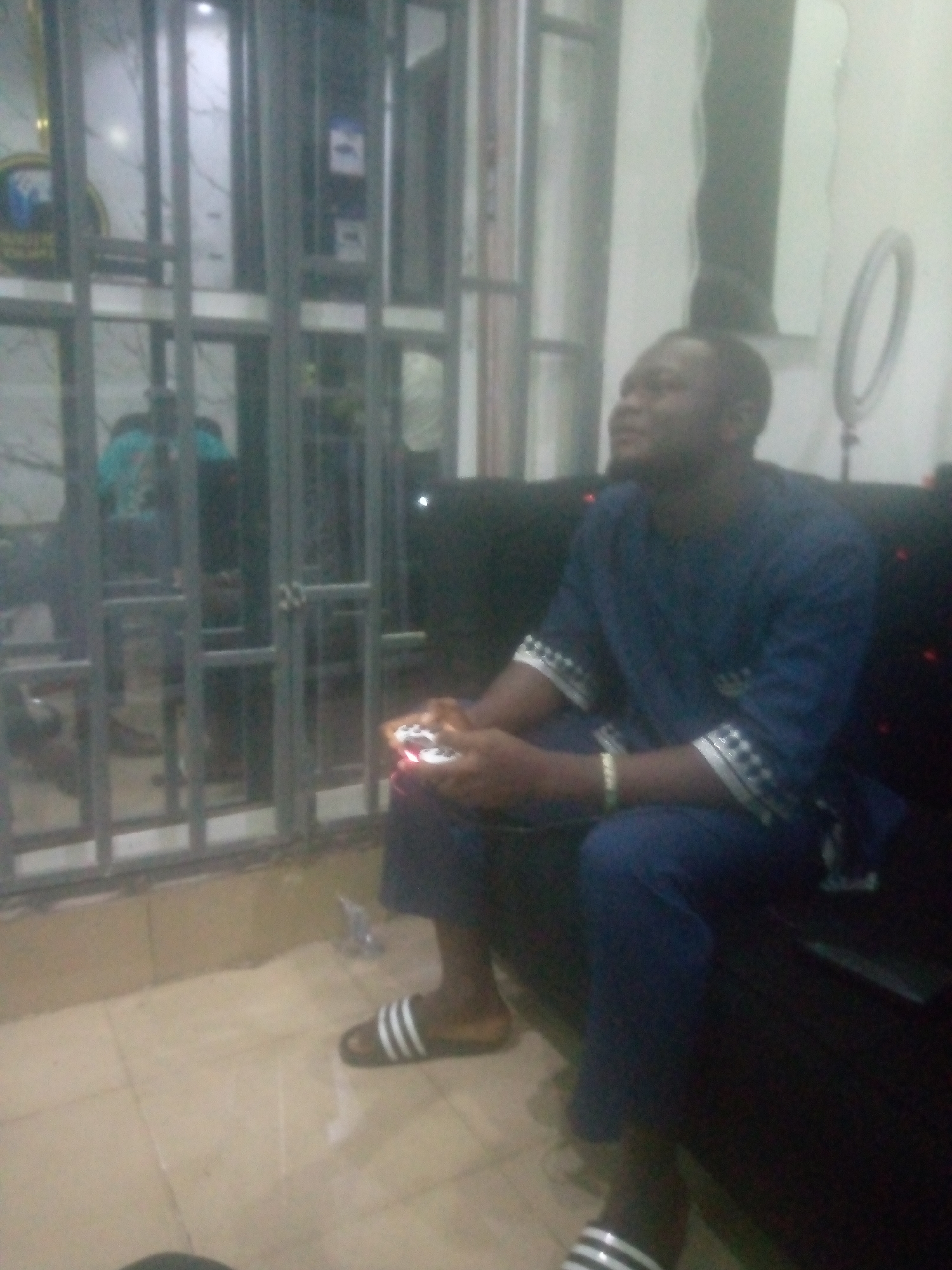 The image depicts an indoor scene in Unknown, Unknown. A male individual, appearing to be of African descent, is seated on a dark, possibly upholstered, couch or bench on the right side of the frame. He is wearing a dark blue tunic-style shirt with patterned white and black cuffs and collar accents, along with matching dark blue trousers. On his feet are black and white striped slides, and a light-colored bracelet is visible on his left wrist. He holds a white electronic device, possibly a gaming controller or smartphone, which emits red and orange light from its surface. His head is turned to the right, and his gaze is directed upwards, suggesting engagement with an unseen screen or activity.
To the left of the individual, a window or glass door secured by grey metal bars is visible. Through the bars, blurry reflections and faint outlines of other figures, one wearing a light blue top, are discernible in the background. A partially visible circular golden logo with blue elements is also present behind the bars on the far left. The floor is covered with light-colored ceramic tiles. On the white wall to the right of the man, a black circular ring light is mounted on a stand, and above it, a tall rectangular mirror or panel is affixed. The scene is dimly lit, illuminated by artificial light sources, contributing to the overall low resolution and blurriness of the image.