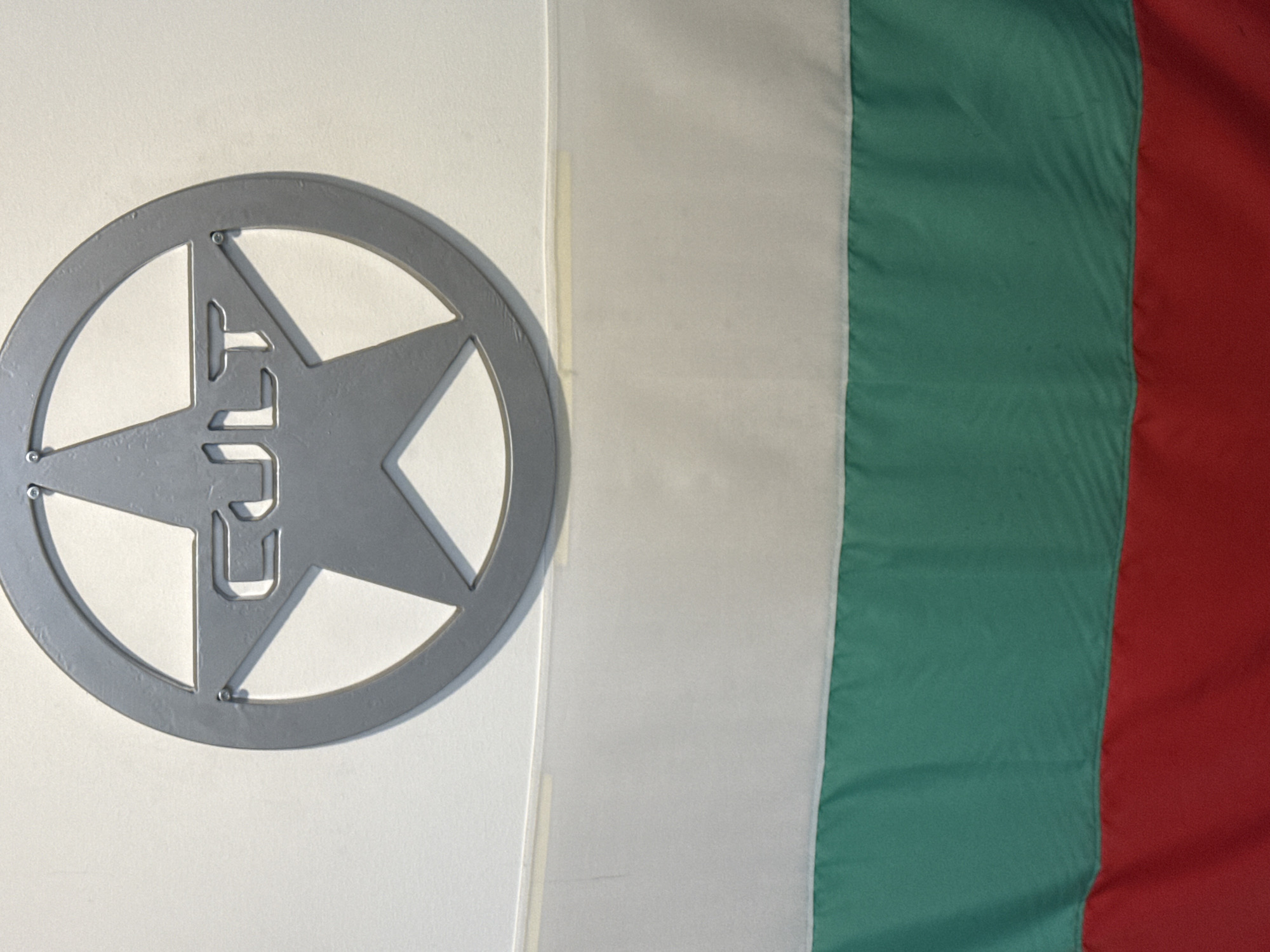 The image captures a static display featuring a metallic emblem and a tricolor flag.
On the left side of the frame, a gray metallic emblem is mounted on a plain white surface. The emblem is circular, with a five-pointed star cut out from its center. Within this star, the word "CULT" is also cut out, creating negative space letters. Small, visible fasteners, possibly rivets or screws, attach parts of the emblem. The emblem casts a faint shadow on the white background, indicating it is slightly raised.
To the right, a fabric flag with vertical stripes is displayed. The colors of the flag, from left to right, are white, green, and red. This color sequence and orientation identify it as the national flag of Bulgaria, consistent with the given location context of Varna, Bulgaria. The flag exhibits slight creases and folds, typical of fabric material. A subtle vertical line or seam separates the white surface holding the emblem from the edge of the flag.
The scene depicts an indoor setting, likely a display or decoration, with no people or specific activity present. The lighting is uniform, providing clear visibility of both the emblem and the flag. The only visible text is the word "CULT" within the emblem.