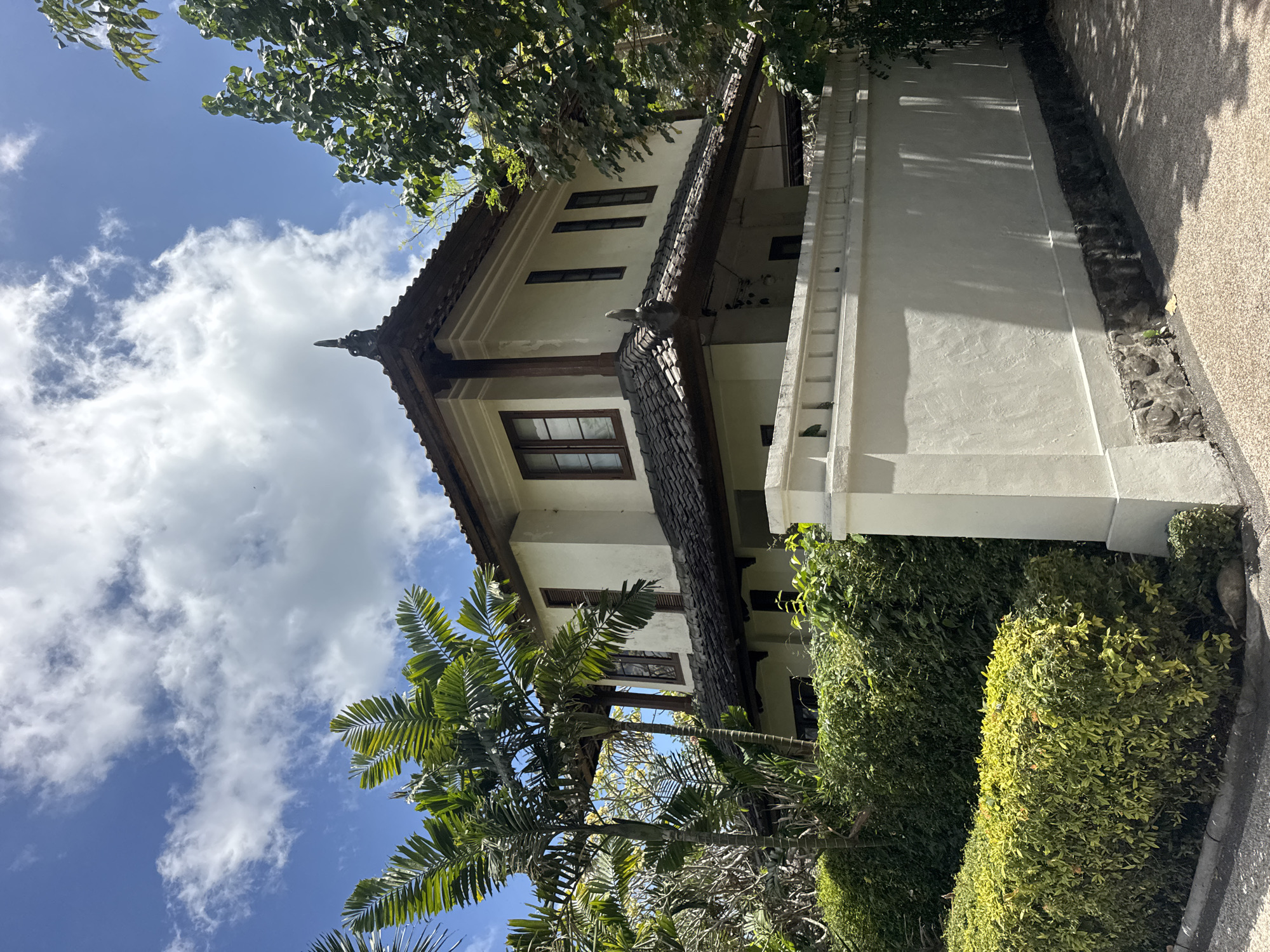 A multi-story cream-colored building with a dark tiled roof and wooden window frames stands amidst lush greenery in Kuta Selatan, Indonesia. The architecture features a prominent decorative spire on the roof and a covered balcony on the second floor. A low, cream-colored wall, topped with decorative elements and accented with stone at its base, encloses the property, separating it from a paved pathway. Several palm trees and dense green bushes surround the building, casting shadows on the wall and ground. The sky above is a vibrant blue, dotted with fluffy white clouds. The scene is illuminated by bright daylight, suggesting it is daytime.