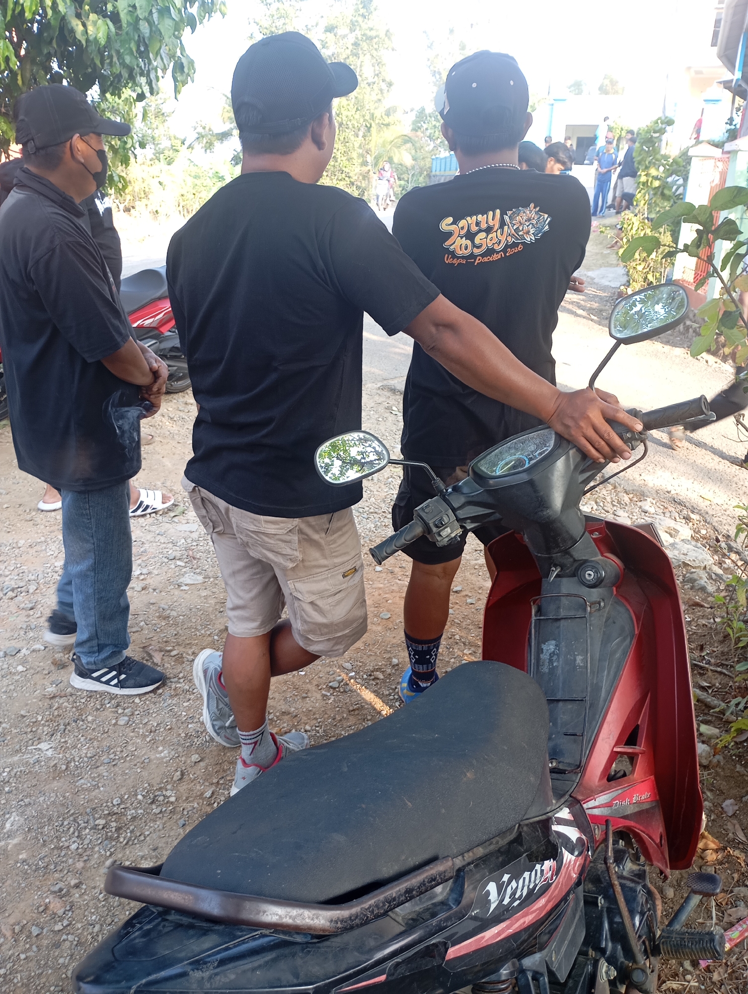 In Ketepung, Indonesia, three men stand near a red motorcycle on a gravelly path. The man on the left wears a black t-shirt, jeans, and a black baseball cap, with a black face mask. The middle man, facing away, is in a black t-shirt, tan cargo shorts, and a black baseball cap. He is holding a cigarette with smoke rising. The third man, also facing away, wears a black t-shirt with a "Sorry to Say Vespa-pacitan 2020" graphic, and black shorts with blue and white patterned socks. His hand rests on the motorcycle's handlebars. The background shows trees and buildings, suggesting a residential area.