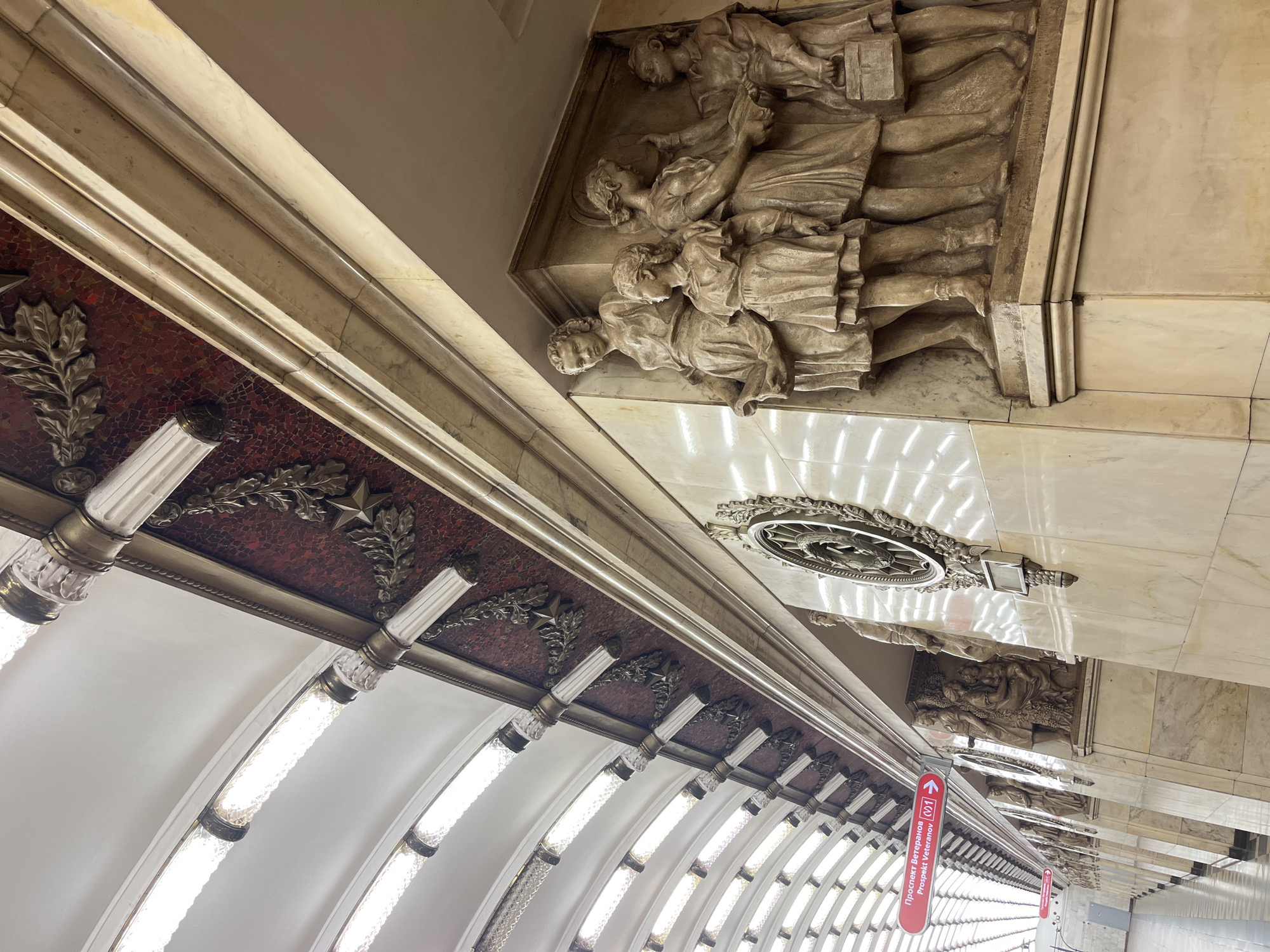 This is an interior shot of the Prospekt Veteranov station on Line 1 of the Saint Petersburg Metro, Russia. The view is an upward angle toward the ceiling. The ceiling is decorated with bas-relief sculptures. The lower portion of the image shows a series of curved, illuminated light fixtures. A sign indicating "Prospekt Veteranov (L1)" is visible. The lighting is bright. The architecture has a classical and ornate style.