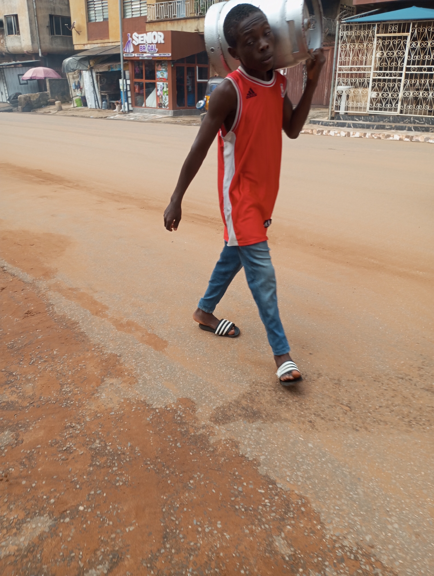 A young Black male, wearing a red sleeveless Adidas shirt and blue jeans, walks on a dusty, unpaved road in Onitsha, Nigeria, carrying a large, silver propane tank on his right shoulder. The tank appears to be empty. In the background, a store with a sign that reads "SENIOR BRIDAL" is visible, along with other buildings and a pink umbrella. The road is a mix of reddish-brown dirt and grey gravel, with some patches of dampness. The scene suggests a typical day in a bustling Nigerian city.