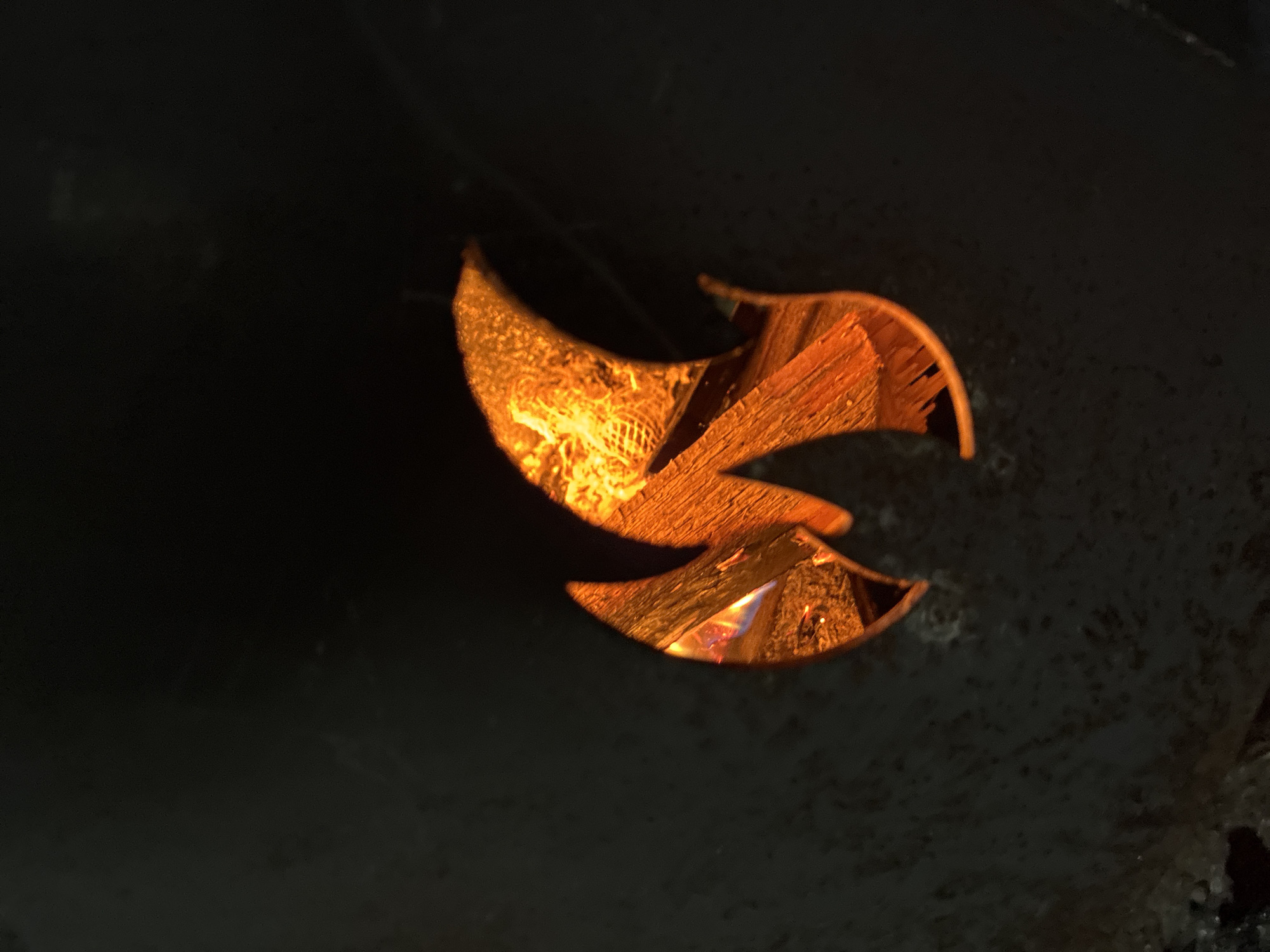 The image depicts a dark, abstract scene with a stylized flame shape in the center, rendered in vibrant orange and yellow hues. The flame is depicted with multiple curved, pointed sections, suggesting movement and intensity. The texture within the flame suggests a granular or splintered material, possibly wood or rock, illuminated from behind. The background is predominantly black, creating a stark contrast that emphasizes the fiery element. There are no discernible people, animals, or identifiable landmarks of Totskoye, Russia in the image. The overall impression is one of abstract art or a symbolic representation of fire, with no specific environmental context provided.