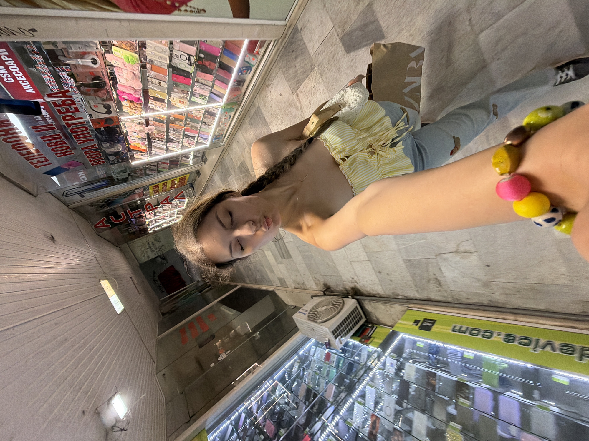 The image depicts a young woman taking a selfie in a narrow, brightly lit commercial passageway, likely a shopping arcade, in Burgas, Bulgaria. The setting is characterized by rows of retail displays filled with phone cases and accessories on both sides of the corridor. The floor is made of light-colored tiles, and the area is illuminated by numerous artificial lights, including LED strips within the display cases.
The woman, positioned towards the center of the frame, has long, dark hair styled in a braid over one shoulder. She is wearing a yellow and white striped strapless top with a tie-front detail and light-colored jeans. A colorful, beaded bracelet adorns her left wrist. Her eyes are closed, and her lips are slightly pursed, suggesting a posed, serene, or playful expression. She holds a light brown paper shopping bag from "ZARA" under her left arm.
Visible text on the shop signs and displays, primarily in Cyrillic, includes "АКСЕССУАРЫ" (Accessories), "ТЕЛЕФОНЫ" (Phones), and phrases like "НОВЫЙ И БУ..." (New and used) and the Bulgarian "ВТОРА УПОТРЕБА" (Second-hand). The abbreviation "GSM" is also seen, referring to mobile phones. On the bottom right, the partial text "device.com" is visible, rotated. An outdoor air conditioning unit is mounted on the wall to the right of the woman.
