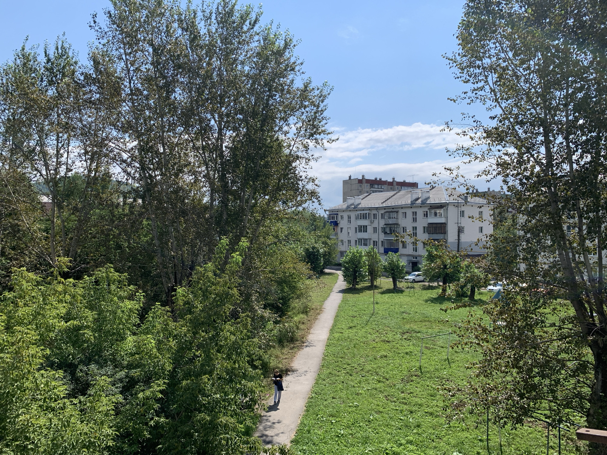 In Asha, Russia, a woman walks on a paved path through a grassy area lined with trees. She is wearing dark clothing and appears to be looking at something she is holding. In the background, a multi-story apartment building with a light-colored facade stands under a blue sky with scattered white clouds. The scene is illuminated by daylight, suggesting it is daytime.