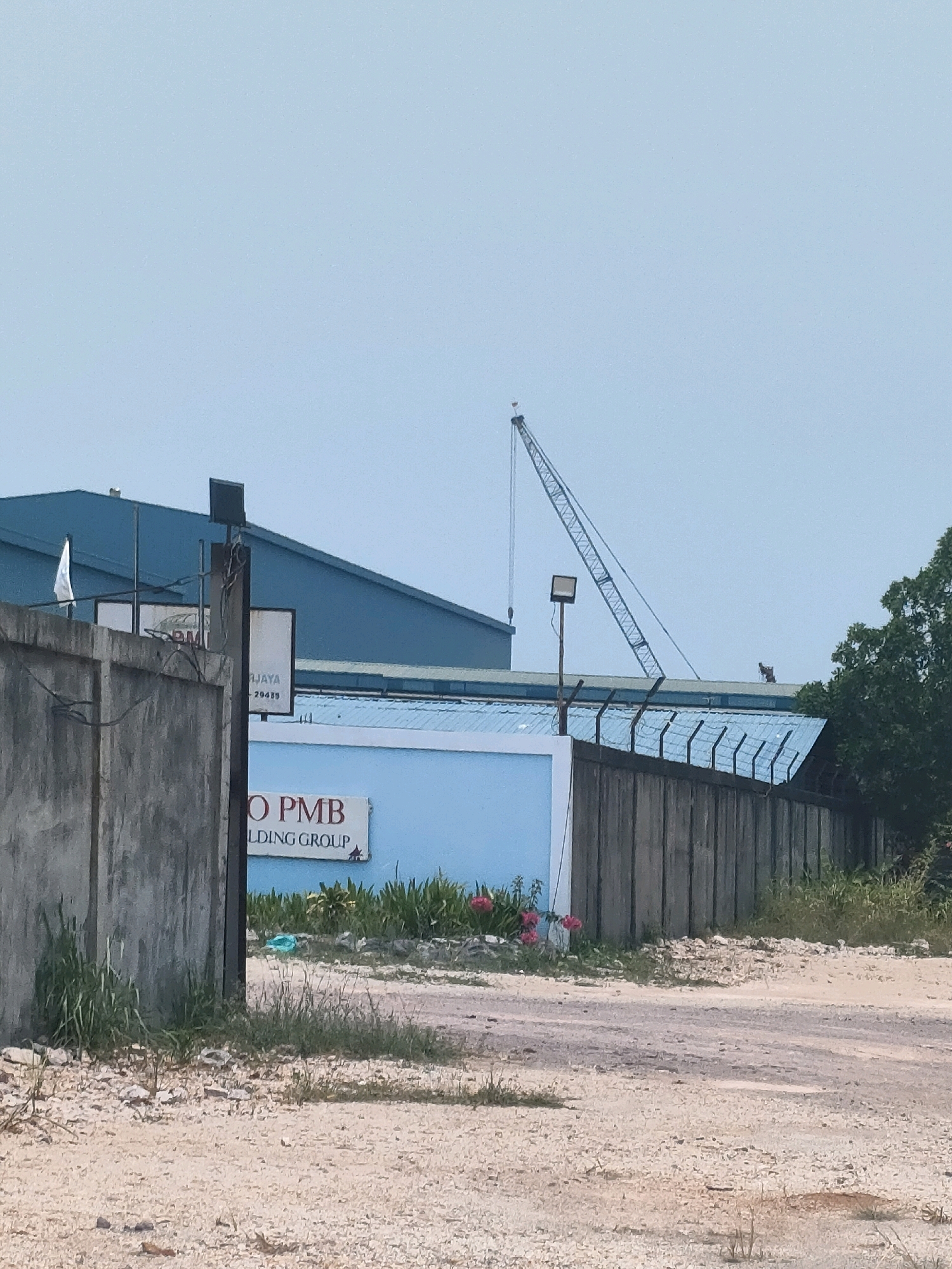 The image shows an industrial scene in Batam, Indonesia. In the foreground, there is a dirt road with scattered rocks and vegetation. A weathered concrete wall runs along the left side of the frame, and beyond it, a large blue industrial building is visible. To the right, a wooden fence topped with barbed wire encloses the property. A lighter blue building with a sign that reads "O PMB LDING GROUP" is situated behind the fence. In the background, a tall construction crane with a lattice boom extends towards the sky, which is overcast. Several trees are visible on the right edge of the frame. The overall scene suggests an active construction or industrial site.