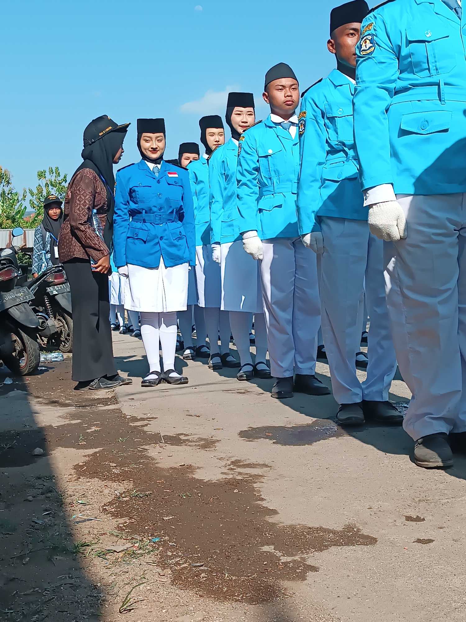 In Imogiri, Indonesia, a group of young people in matching blue uniforms and black hats stand in formation outdoors. The uniforms consist of blue jackets with white collars and ties, white skirts or pants, and white socks. Some individuals are wearing white gloves. To the left, two adult women, one in a brown patterned garment and hijab, and another in a dark hijab and a blue and white patterned shirt, are present. A motorcycle is parked on the left side of the image. The ground is paved and appears wet in places, with some scattered debris. The background shows a clear blue sky with a hint of a moon, and some greenery and structures can be seen in the distance.
