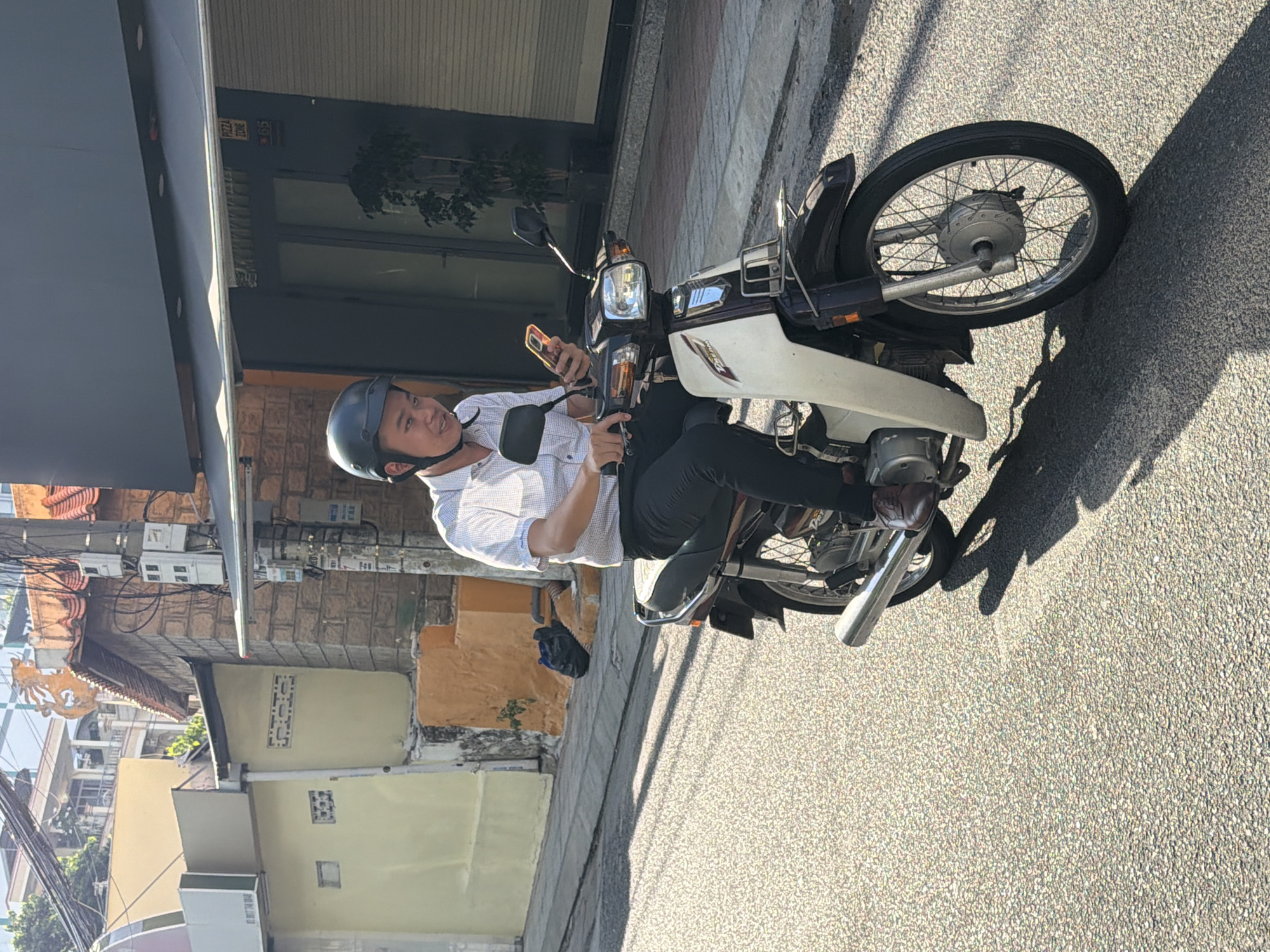 In Nha Trang, Vietnam, a young man is riding a dark brown and white scooter. He is wearing a light blue and white checkered shirt, dark pants, brown shoes, and a dark helmet. He is looking down at his orange smartphone, which he holds in his right hand while steering with his left. The scooter is moving on a paved street with sunlight casting shadows. In the background, there are buildings, including one with a yellow facade and a dark awning with a "PIZZA ZONE" sign and the number "65." There are also utility poles with wires and electrical boxes. A black plastic bag lies on the orange ground in front of a building.