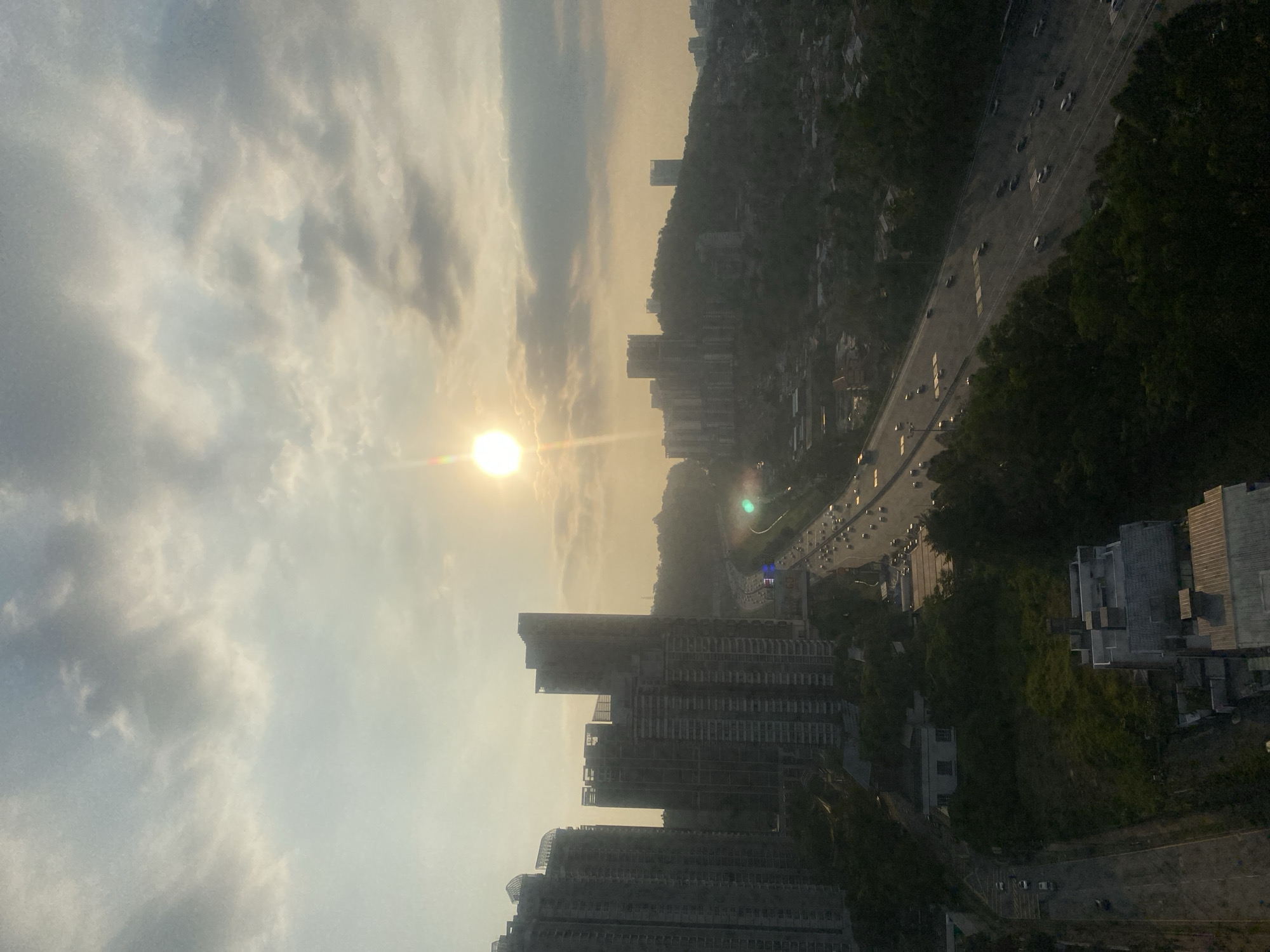 The image captures a panoramic view of Kuala Lumpur, Malaysia, during what appears to be either sunrise or sunset, given the low angle of the sun and the warm, hazy light illuminating the sky. In the foreground, on the left side, several tall, modern high-rise buildings are visible, some of which appear to be under construction, indicated by exposed structural elements and unfinished facades.
The midground features a multi-lane highway congested with traffic, with cars and trucks moving in both directions. The highway cuts through a landscape characterized by rolling hills covered in dense green foliage, dotted with more buildings, including lower-rise residential structures and commercial establishments. Some of these buildings are clustered on the hillsides, while others are situated closer to the highway.
In the distance, the urban sprawl of Kuala Lumpur extends across the horizon, with more buildings and infrastructure visible under the soft light. The sky is partly cloudy, with a mix of light and darker gray clouds, through which the sun shines brightly, casting a golden glow and creating lens flare effects. The overall atmosphere is one of a busy, developing city bathed in natural light.