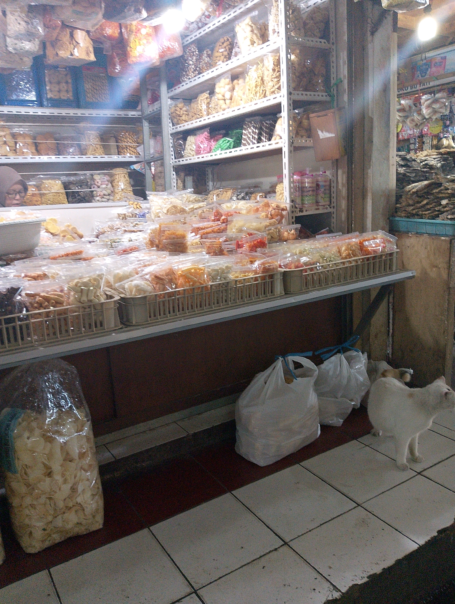 A street-level view captures an outdoor market stall in Lembang, Indonesia, displaying an assortment of packaged snacks and dried goods. The stall is filled with shelves stacked high with various food items, mostly in transparent plastic packaging, including what appear to be crackers, chips, and other dried treats. A person, wearing glasses and a headscarf, is partially visible in the upper left quadrant, appearing to examine or interact with the merchandise. In the foreground, a large, clear plastic bag is filled with what looks like light-colored, thinly sliced snacks. To the right, two white plastic shopping bags are placed on the ground, and a white cat stands near them, looking upwards and to the right. The flooring is tiled with a pattern of squares. The overall scene suggests a bustling market environment focused on food sales.