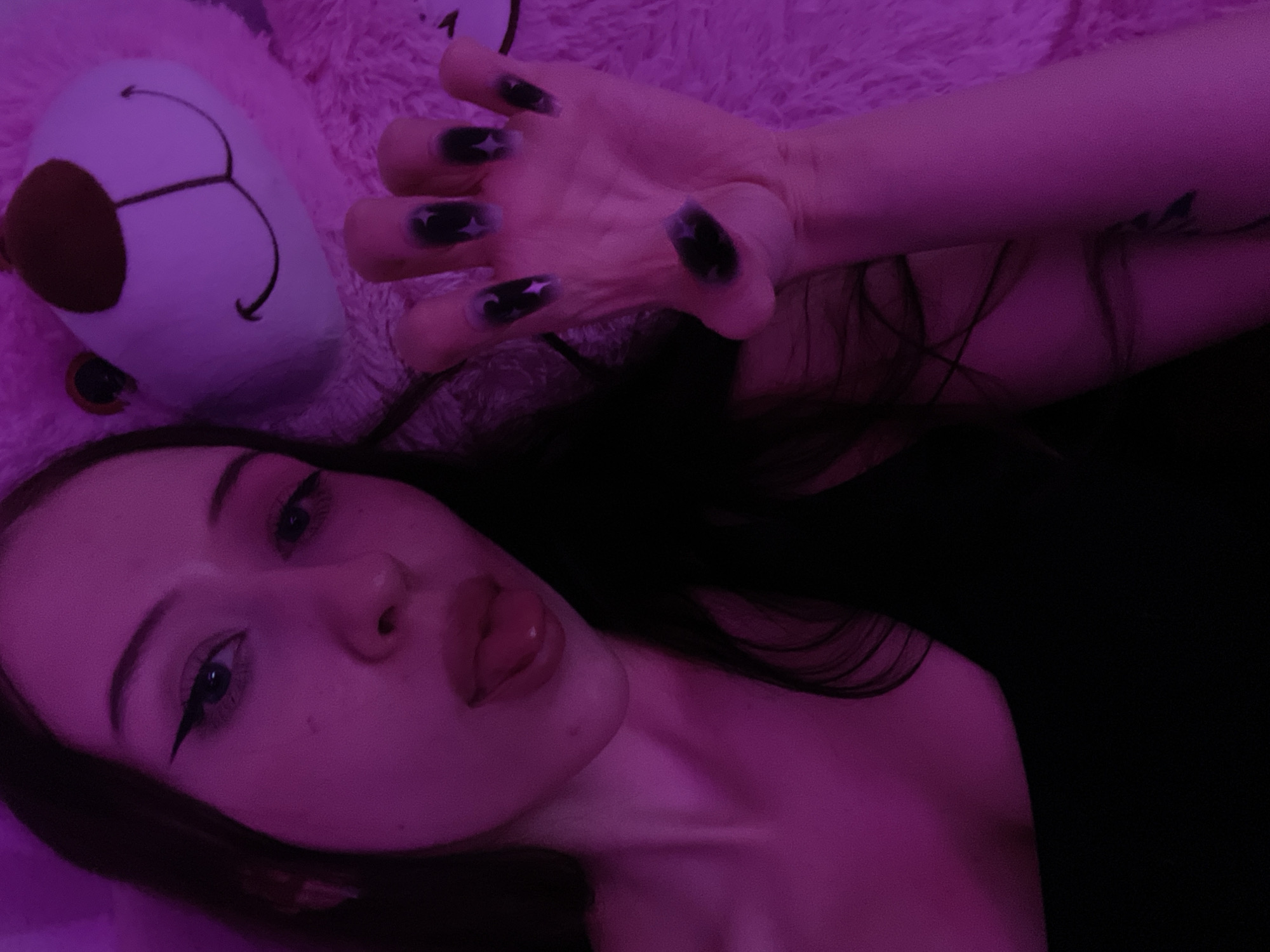 The image shows a young woman with long dark hair, wearing black clothing. She has dramatic winged eyeliner and her tongue is sticking out. Her right hand is raised with long, dark, pointed nails, each decorated with a white star. She is posing in front of a large, fluffy pink teddy bear. The scene is illuminated by purple lighting, creating a moody atmosphere. The image does not contain any information that would allow for the determination of its location in Votkinsk, Russia.