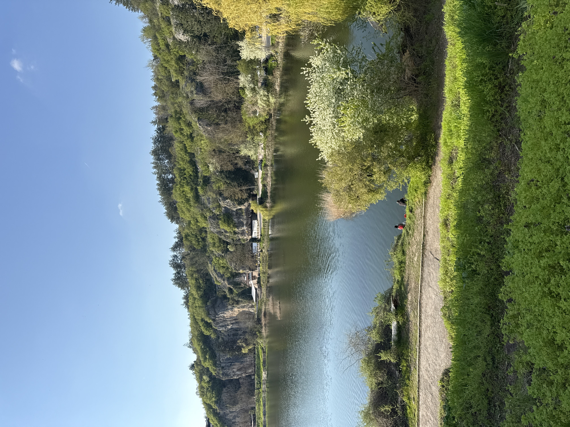 The image depicts a landscape in the city of Pleven, Bulgaria, featuring a river or lake running parallel to a cliff face covered in trees. Along the edge of the water is a narrow path, bordered by green foliage. Two people, one wearing a red garment, are visible at the edge of the water. The sky is clear blue. Buildings or structures are visible further up the cliff face.