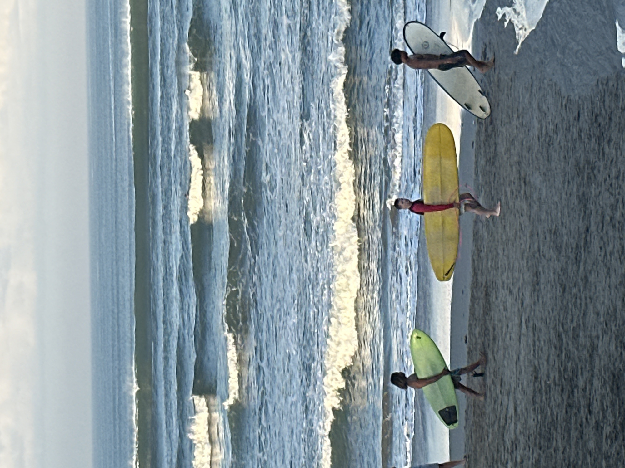 Three individuals are walking on a sandy beach along the coast of North Kuta, Indonesia, carrying surfboards. The person on the left carries a lime green surfboard. The person in the middle carries a bright yellow surfboard. The person on the right carries a white surfboard with black fins. The ocean is in the background, with several lines of waves breaking. The sky is overcast.