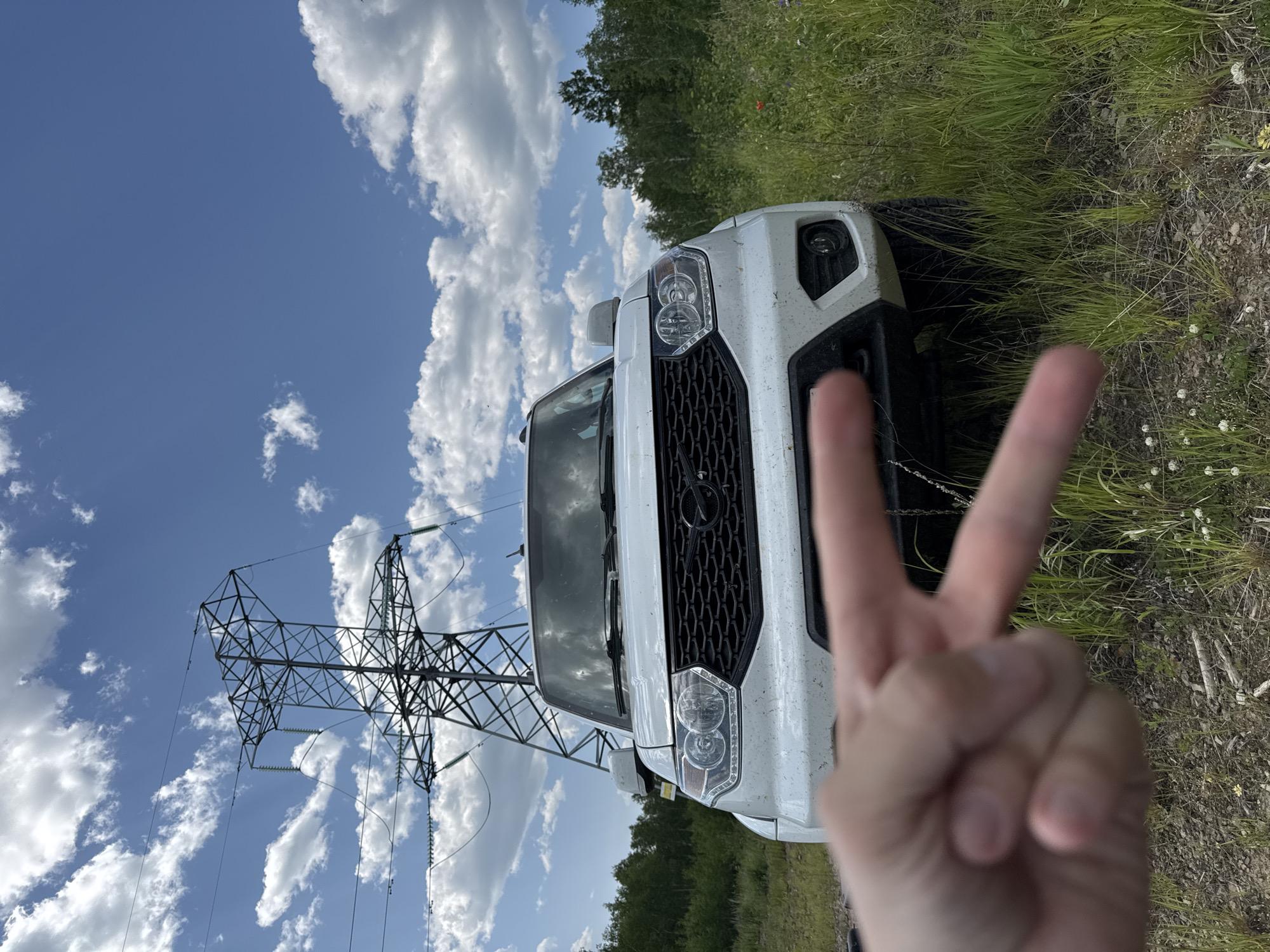 A white SUV is parked in a grassy field with a tall electrical tower in the background. The sky is blue with scattered white clouds. A hand is in the foreground making a peace sign. The car appears to be dusty, suggesting it has been driven off-road. The overall scene depicts an outdoor recreational setting. The location is identified as Skovorodino, Russia.