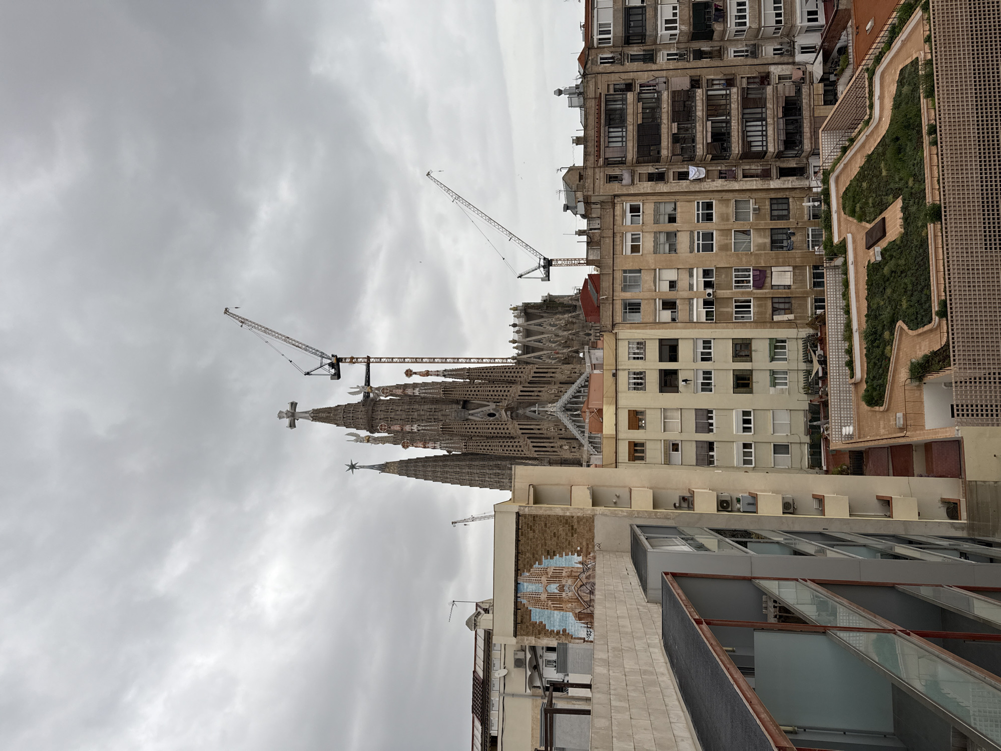 The photograph showcases an upward angle view of a skyline in Barcelona, Spain, dominated by the Sagrada Família, a large unfinished Roman Catholic minor basilica. Two cranes hover beside the cathedral, signifying ongoing construction. The architectural diversity of the surrounding buildings is apparent, with structures varying in height, color, and material. Some are older, multi-story apartments with traditional windows and facades, while others appear more contemporary with glass panels and modernized designs. A building on the right is adorned with a terrace garden on its roof. Overcast skies fill the top half of the image, casting a muted tone over the scene. No people are visible in the frame.