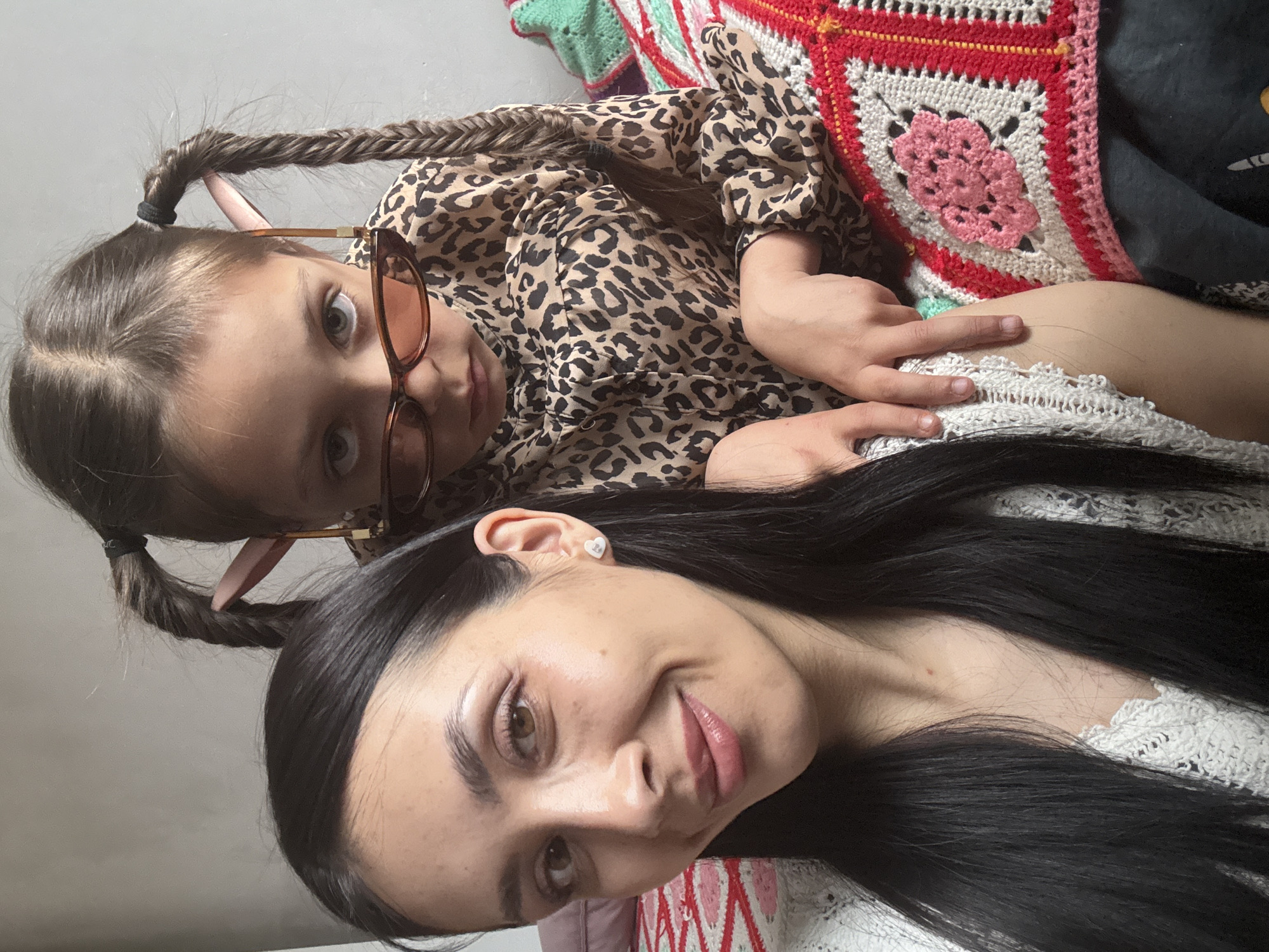 Two individuals, likely a mother and daughter, are posing for a selfie indoors. The mother, with long black hair and light skin, is in the foreground. She wears a light-colored garment with a lace-like texture. The daughter, who has braided hair and wears glasses, is positioned behind and slightly above the mother. She wears a leopard-print top and her arm and hand are visible. A colorful crocheted blanket serves as a backdrop. The location is not evident in the picture, so it cannot be determined if the scene is in Votkinsk, Russia.