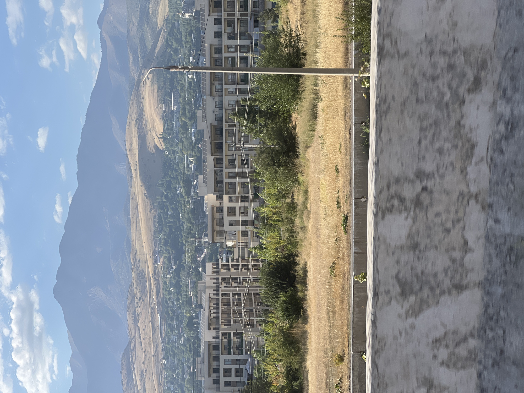 A wide shot captures a vast mountainous landscape behind a residential area in Chinarkent, Uzbekistan. In the foreground, a grey concrete barrier runs across the bottom of the frame. Beyond this barrier, a strip of dry, golden grass is visible, bordered by a low concrete curb. Behind the grass, a line of lush green trees forms a dense foliage line.
Dominating the mid-ground is a large, modern, multi-story apartment complex. The building features a beige facade with numerous balconies and rows of windows. Several utility poles and power lines are scattered in front of the building and within the trees.
Further in the background, a sprawling town or city extends across rolling hills. The terrain gradually slopes upwards, leading to a range of imposing mountains in the distance. The mountains are muted in color, appearing hazy blue and grey against a bright, partly cloudy sky. The overall scene suggests a sunny day with a clear atmosphere.