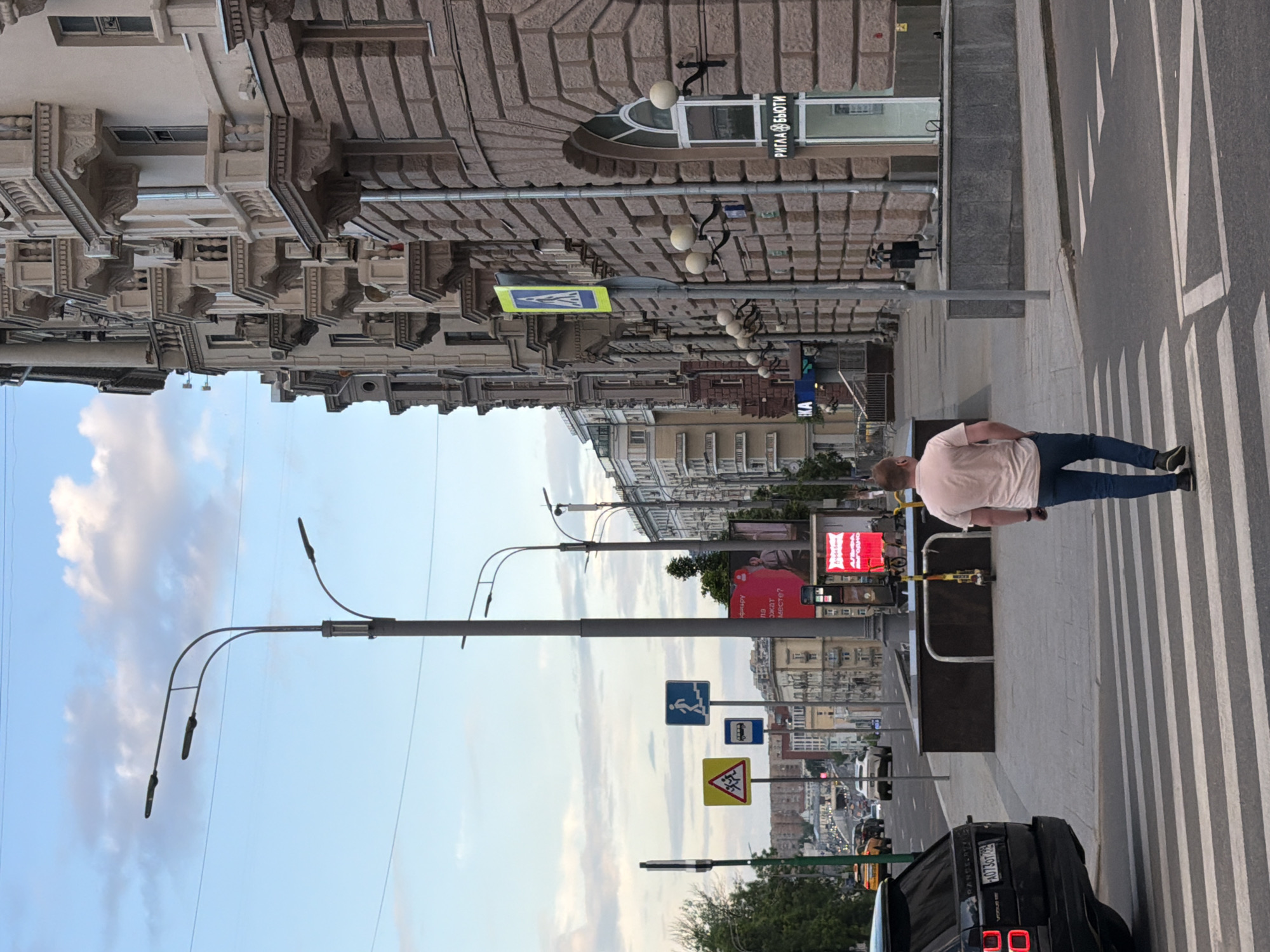 The image shows a street scene in an urban area, specifically a crosswalk. A man in a pink shirt and jeans is walking across the crosswalk. A black Range Rover with the license plate "A073OT 799" is visible. Traffic signs and streetlights are present along the street. Buildings are visible on both sides of the street, showcasing varied architectural styles. A sign with text in Russian ("РИГЛА @ БЬЮТИ") is noticeable on one of the buildings. The sky is partially cloudy.