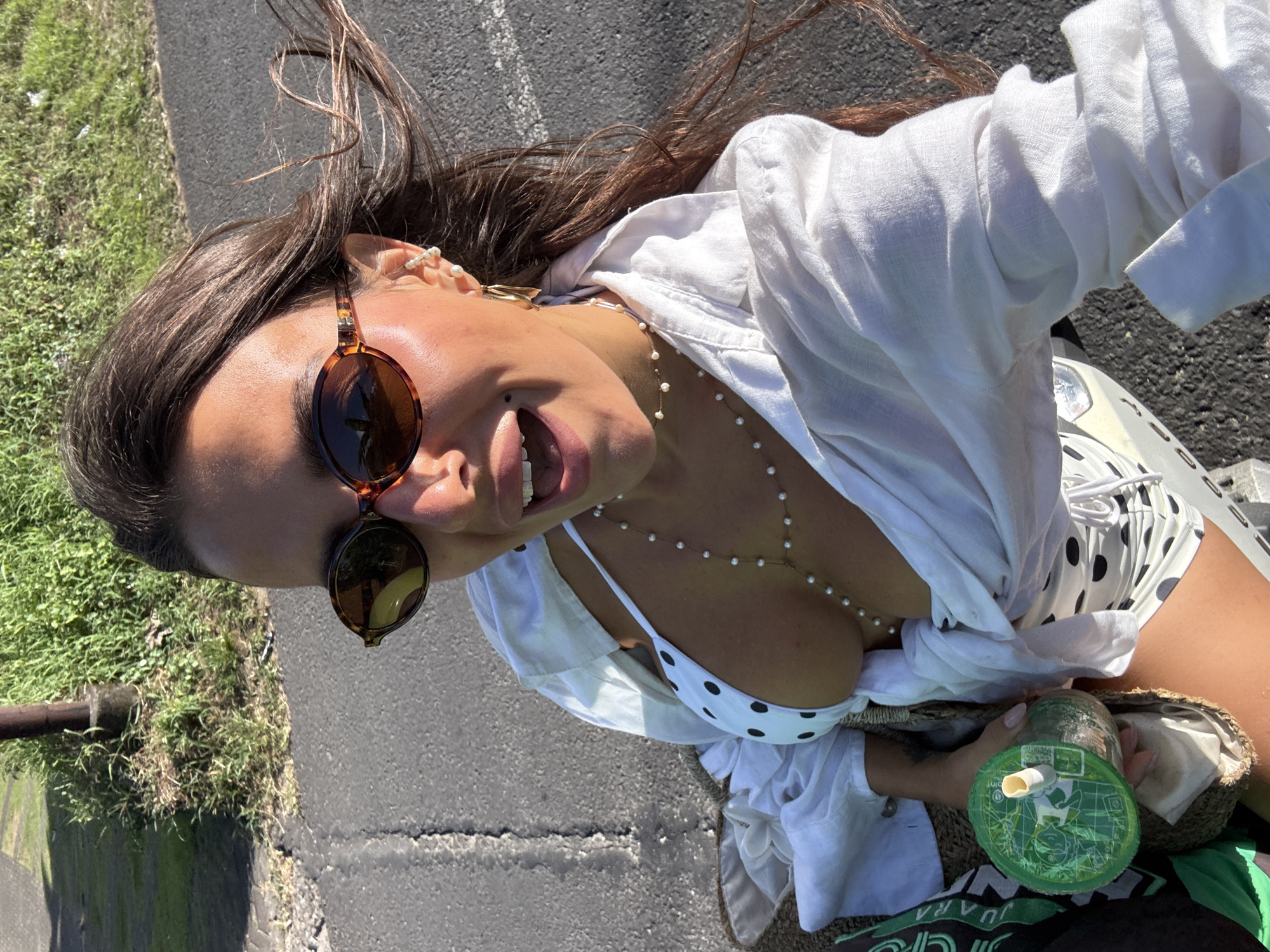 A woman is posing for a photo on a paved road. She is wearing sunglasses pushed up on her head, a white linen shirt over a polka-dot bikini top, and a pearl necklace. She is holding a drink in a straw bag. The background includes green grass and a roadside ditch. The location is likely near a beach or coastal area in Kuta Selatan, Indonesia.