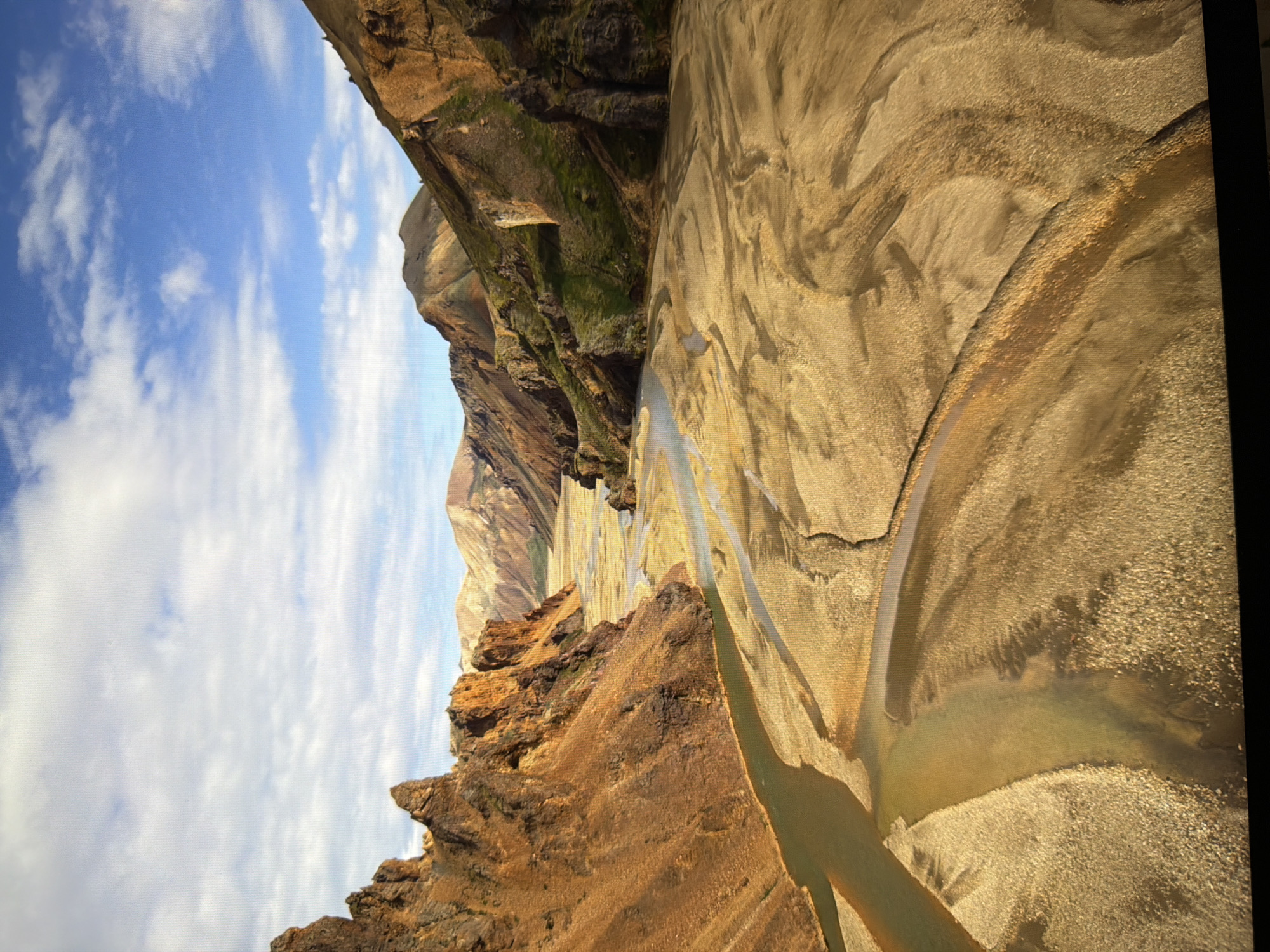 This is a wide-angle, aerial photograph of a rugged, mountainous landscape under a partially cloudy blue sky. A river with multiple channels winds through a valley filled with light-colored sand and gravel. Steep, ochre-colored cliffs rise on the left side of the valley, with patches of green vegetation on the slopes of the mountains in the distance. The overall impression is one of a wild, remote, and arid environment. The image does not depict any elements related to the city of Kyiv, Ukraine.