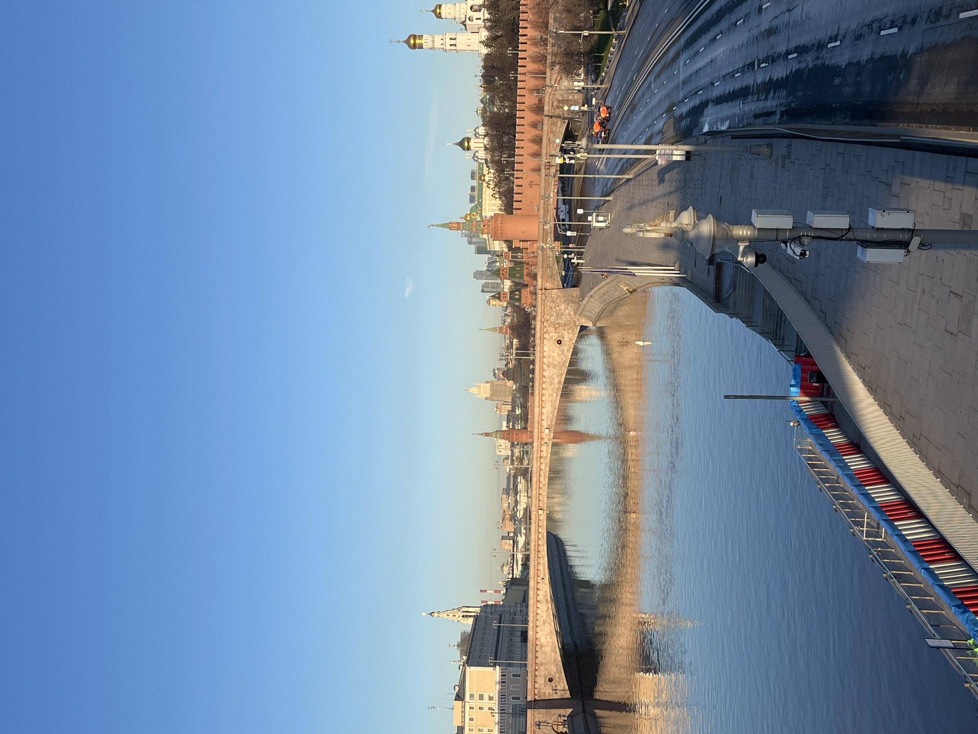 The image presents a view of the Moscow Kremlin reflected in the Moskva River, taken from a vantage point along the embankment. The perspective is from near the riverbank, offering a wide view across the water towards the Kremlin's walls and towers. The Kremlin, including its iconic cathedrals with golden domes, and the fortress wall, is clearly visible. The reflection in the water mirrors the skyline, providing a symmetrical effect. The river's surface is relatively calm, enhancing the clarity of the reflection. Along the river's edge, some structures are visible. The sky is clear and blue, contributing to the overall clarity of the scene. The image captures a moment during daylight, possibly early morning or late afternoon, given the quality of light. The roadway and sidewalk along the river near the Kremlin are also observable.