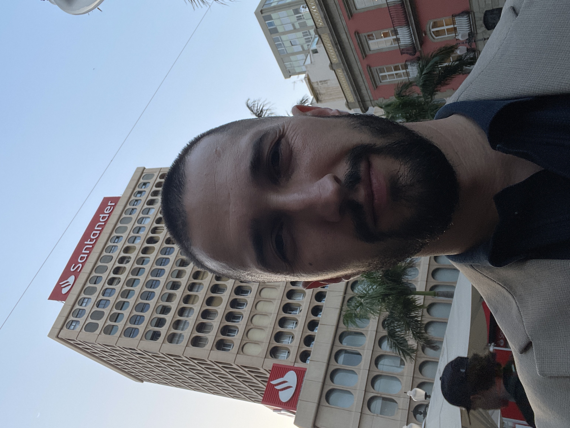 The image presents a close-up, low-angle perspective of a man in an urban setting. The man, with a beard and short dark hair, is positioned in the foreground, looking towards the camera with a neutral expression. He is wearing a blue shirt layered under a light-colored jacket.
Behind the man, a prominent multi-story building with a unique beige facade featuring numerous oval or pill-shaped windows extends upwards. The red "Santander" text and its characteristic flame-like logo are clearly visible on two different sections of this building, indicating its affiliation with the bank. Several palm trees are interspersed around the base and mid-section of this structure.
In the further background, another building with a reddish facade and balconies is partially visible. The sky is bright and clear, suggesting a daytime scene, possibly late afternoon given the warm light on the man's face. A street light pole and a partially visible individual wearing a cap can also be discerned in the lower right portion of the frame. The overall setting depicts a busy city environment, consistent with Santa Cruz De Tenerife, Spain.