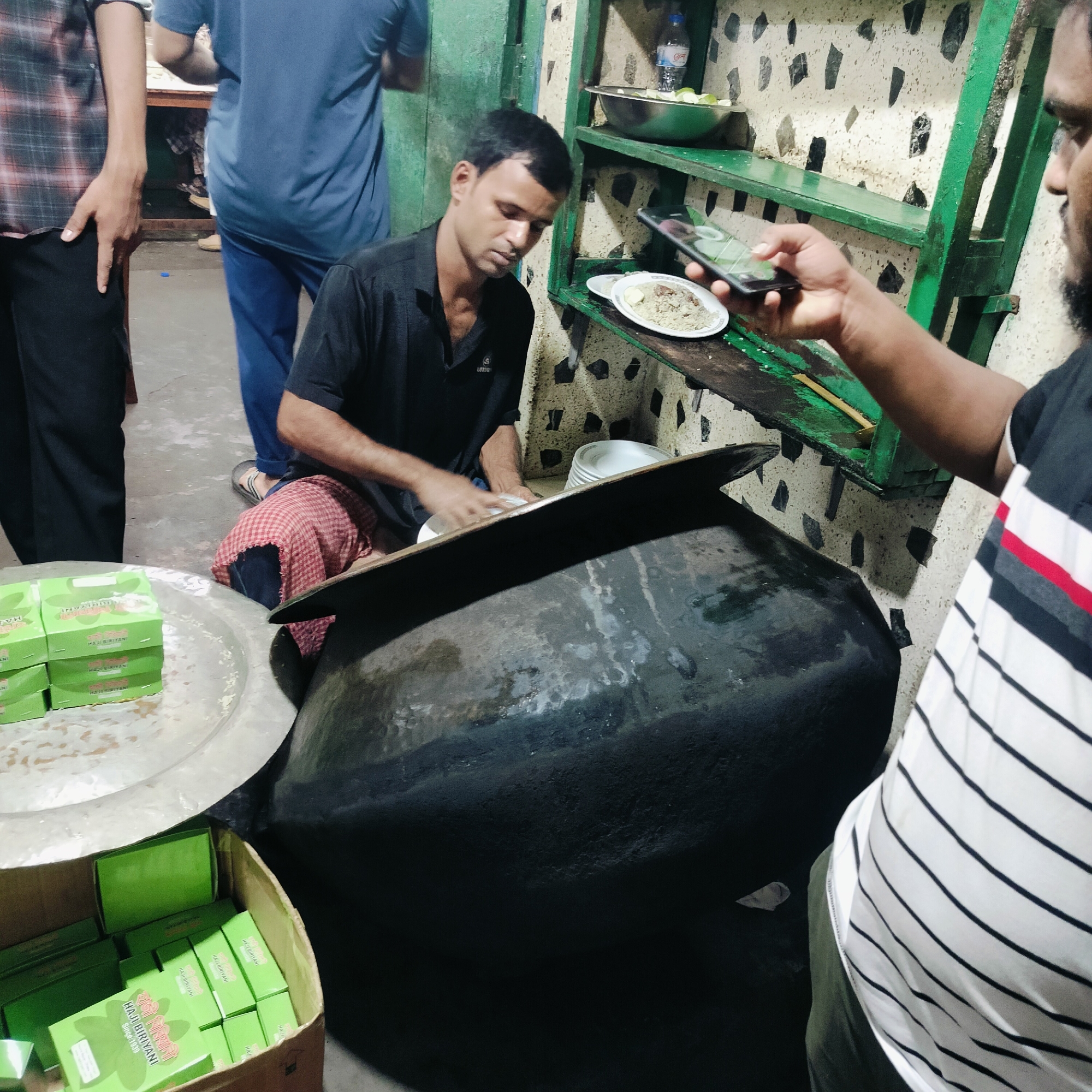 In Dhaka, Bangladesh, a scene unfolds in what appears to be a bustling food establishment, possibly a restaurant or street food stall. The foreground is dominated by a large, dark, metallic pot, likely used for cooking. To the left, several stacks of green boxes, labeled "HAJI BIRYANI Since 1830," are visible, suggesting that biryani is a specialty here.
A man in a black polo shirt is the central figure, actively engaged in preparing or serving food. His hands are busy over a large pot or serving vessel, and he appears to be concentrating on his task. He is wearing a red and white checkered cloth around his waist. To the right, another individual, partially visible, holds a smartphone, possibly taking a photograph or video of the scene. This person is wearing a black and white striped shirt.
In the background, shelves are adorned with various items, including a bowl of green slices, likely limes or similar fruit, and a bottle of water. The walls have a speckled pattern, with dark, irregular shapes against a lighter background. Another person in a blue t-shirt and blue trousers is also present, standing near a counter. The overall environment suggests a busy, operational food service setting.