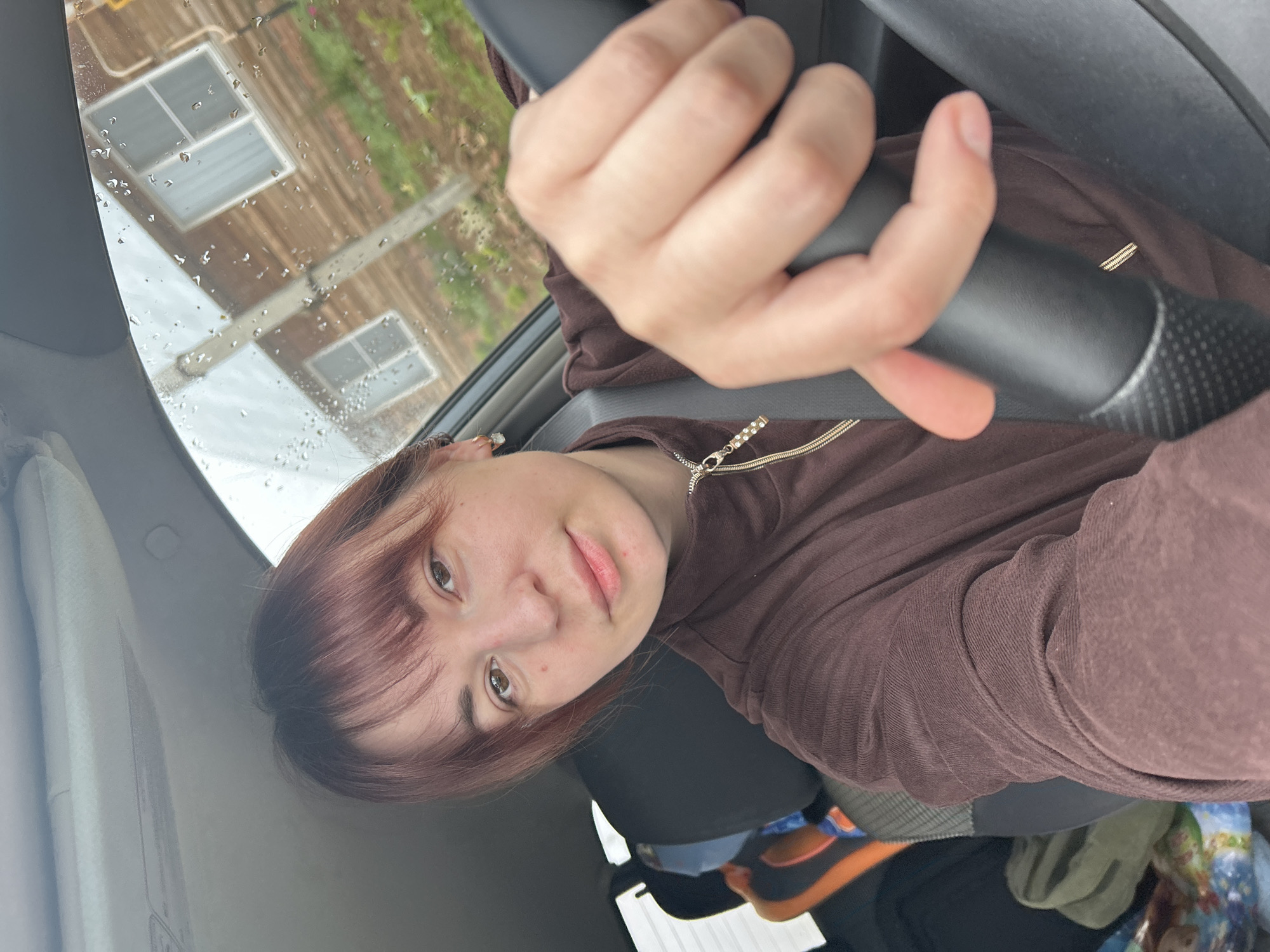A young woman is captured in a selfie taken from the driver's seat of a car. She has reddish-brown hair with bangs, light skin, and brown eyes, looking directly at the camera with a neutral expression. She is wearing a brown hooded jacket with a decorative gold zipper featuring small clear stones. A seatbelt crosses her chest, and her right ear displays a small, sparkly earring. Her hands are positioned on the steering wheel, which has a textured grip.
The setting is inside a car during daytime, with the windshield covered in numerous water droplets, indicating rain or recent rain. Through the wet glass, an exterior scene is visible: a building with two rectangular windows, suggesting a wooden or brick structure, is partially obscured by green foliage in the background. Inside the car, a black car seat and miscellaneous items, including an orange object and green fabric, are visible on the floor. The image is rotated clockwise, presenting a sideways view of the subject. There is no visible text.