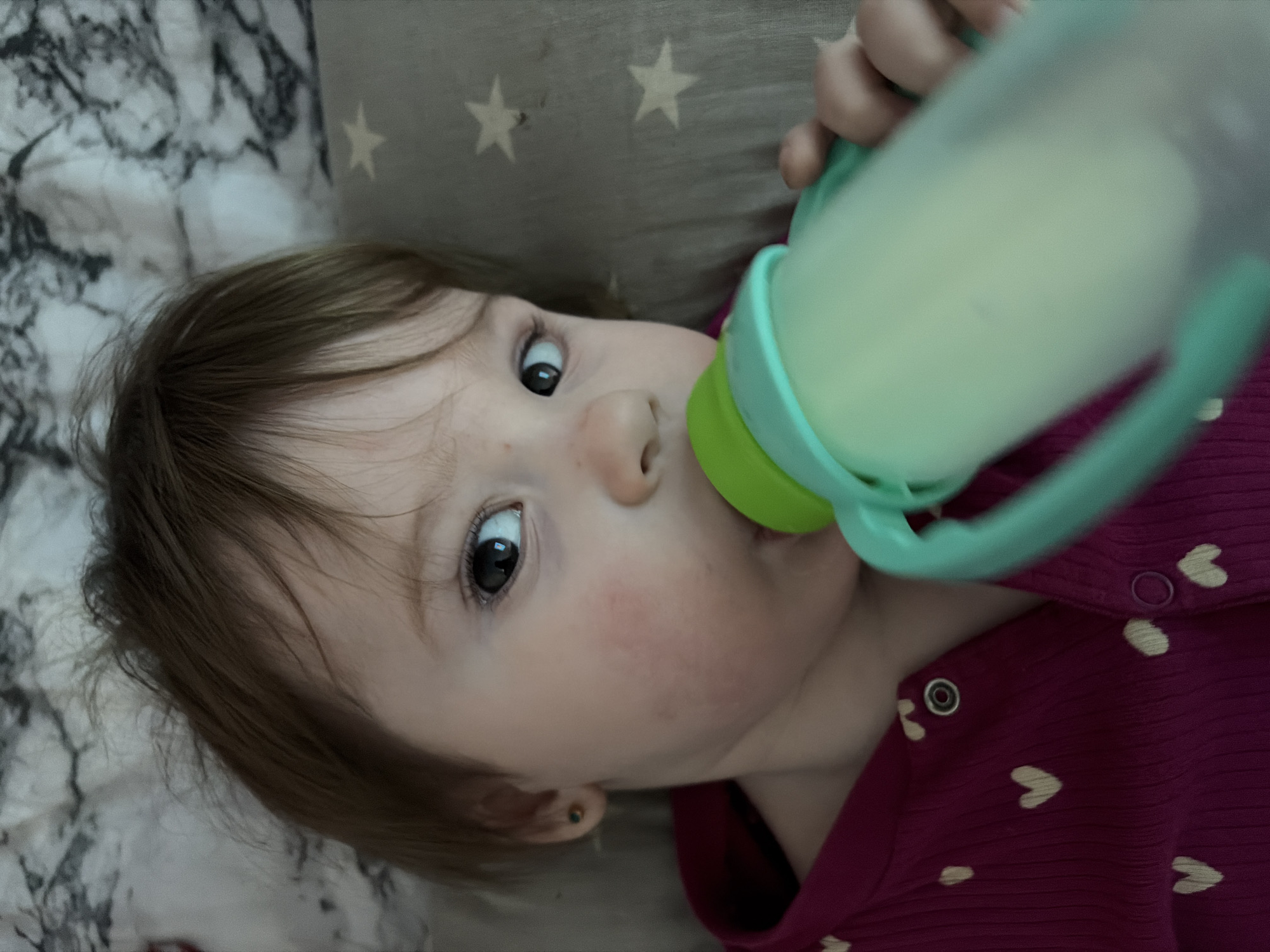 An overhead, slightly angled shot captures a baby or toddler drinking from a bottle while lying down. The baby's face is central and in focus, featuring wide, dark eyes looking directly at the camera. Their light brown hair frames the face, and a slight flush is visible on their cheeks. A small, dark stud earring can be seen in their left earlobe.
The baby is holding a clear baby bottle or sippy cup with bright green and turquoise accents (including handles) up to their mouth. The bottle contains a light, milky liquid, consistent with formula or milk, and is slightly out of focus in the foreground.
The baby is wearing a dark pink or magenta top adorned with small, light-colored heart patterns and snap fasteners on the shoulder. Behind the baby's head, a grey pillow with white star patterns is visible, contributing to a cozy, childlike atmosphere. To the left of the baby, a white and grey marbled pattern, possibly a sheet or blanket, adds texture to the background. The scene suggests an indoor setting, likely in a bed or on a comfortable surface.
