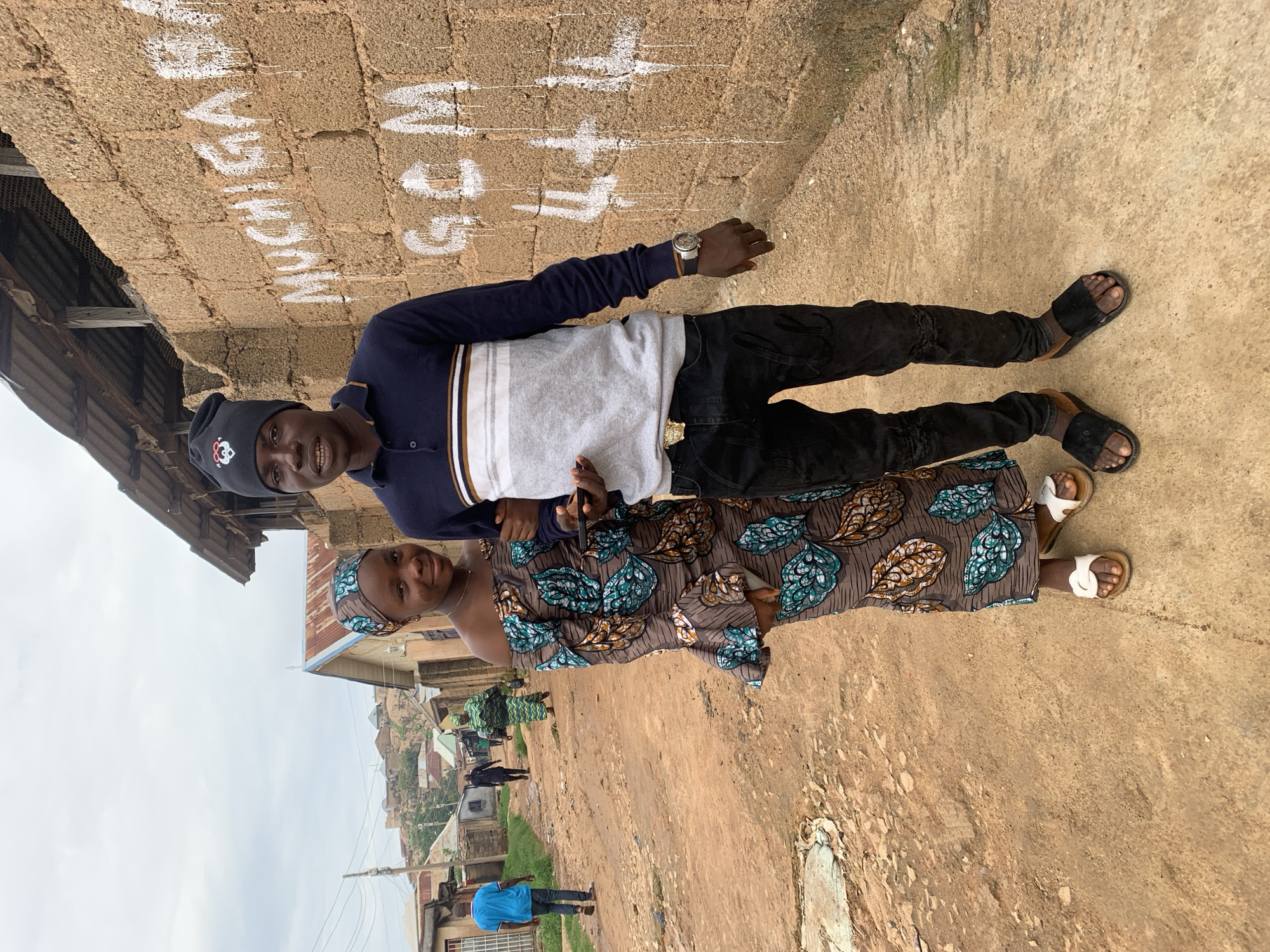A candid full-body photograph captures a young couple standing on a mixed dirt and concrete path in a residential area of Jos, Nigeria. The sky above is overcast and pale gray.
In the foreground, a young woman with dark skin stands on the left, linked arm-in-arm with a young man on the right. She wears a colorful headscarf and a matching off-the-shoulder maxi dress, both patterned with large teal, gold, and brown leaf-like motifs. She has white flat sandals and a small necklace, and holds a dark object in her left hand. She is smiling faintly at the camera. The man, also with dark skin, wears a black beanie with a white logo, a dark blue polo shirt with horizontal white, gray, and gold stripes across the chest, and black ripped jeans with subtle embroidery near the knees. He wears a gold Versace belt buckle, a wristwatch on his left wrist, and black slide sandals. He holds a black smartphone in his right hand and smiles broadly, showing his teeth.
Immediately to their right is a wall constructed of unpainted, rough-textured brown concrete blocks. White graffiti is painted on this wall, reading "MUCHIGABA" (or a similar word, with some fading), followed by "GCM" below it, and then two "U" shapes and three "+" signs (UU+++) with visible paint drips. Above the wall, a structure with a corrugated metal roof is visible.
The ground beneath them is a blend of packed orange-brown dirt and patches of lighter-colored concrete. To the left, a dirt road extends into the background, flanked by various single-story residential buildings made of concrete blocks, some unpainted, others with light-colored finishes. Several individuals are visible walking on the road in the mid-ground: a man in a blue t-shirt walks away on the far left, and further down, a person in a patterned green dress walks towards the foreground, alongside another figure in dark clothing. In the distant background, rolling hills with scattered dwellings are discernible under the cloudy sky.