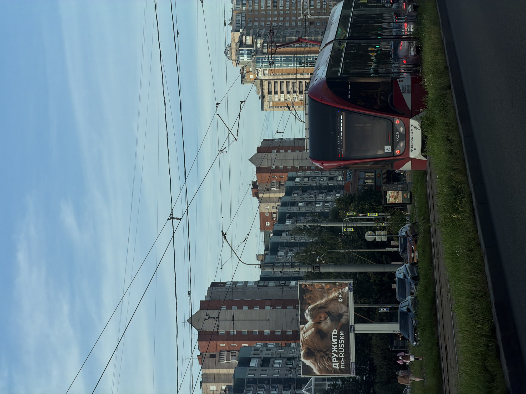 The image captures a street scene in a city, likely during daylight with some cloud cover. In the foreground, a modern red and white tram is visible on tracks, with its front displaying route information and a number 7848. To the left of the tram, several cars are parked on the side of the road, behind a patch of lush green grass. Further back, a large billboard features an advertisement with an image of a bear and a young girl. Beyond the billboard and cars, a row of residential buildings, some with brick facades and others with gray and green exteriors, stretch across the middle ground. In the background, taller, more modern buildings with glass facades are prominent. Overhead, a complex network of power lines crisscrosses the sky, typical of urban infrastructure. A few people are seen walking on the sidewalk near the cars and billboard. The overall impression is an active urban environment with a mix of older and newer architectural styles.