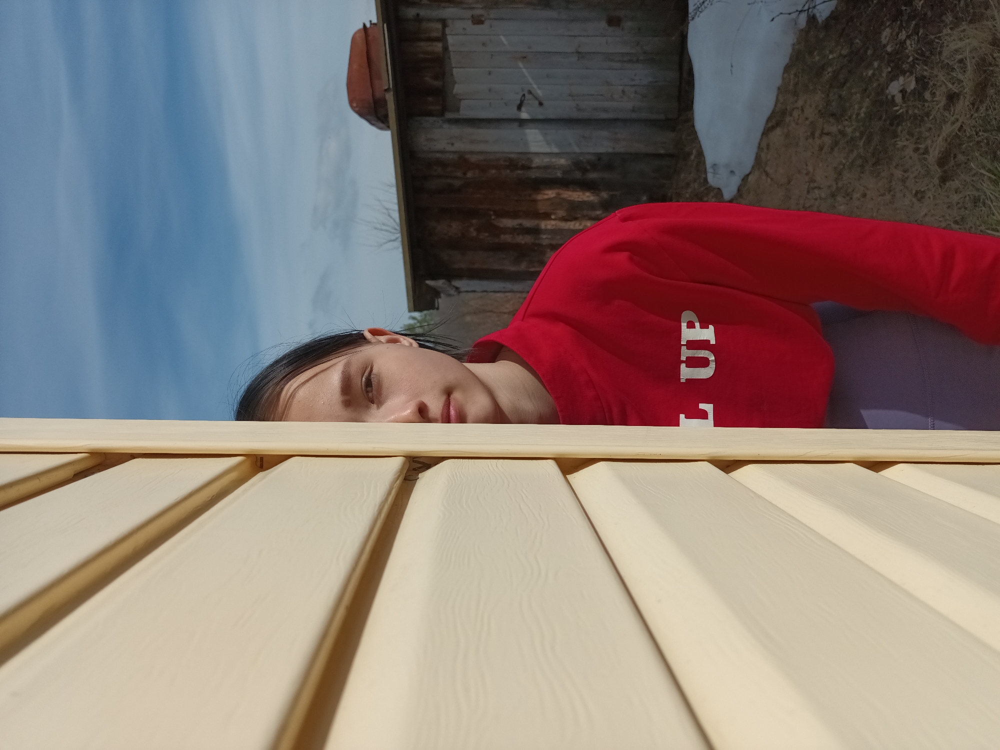 A woman with dark hair and fair skin is partially visible behind a light yellow wooden structure with linear patterns. She is wearing a red sweater with white lettering, and possibly purple clothing underneath. The background includes a blue sky with scattered clouds, a weathered wooden building with a rusty-colored object on top, and patches of snow or light-colored debris on the ground. The image was taken outdoors, and potentially in the city of Ольховка, Russia.