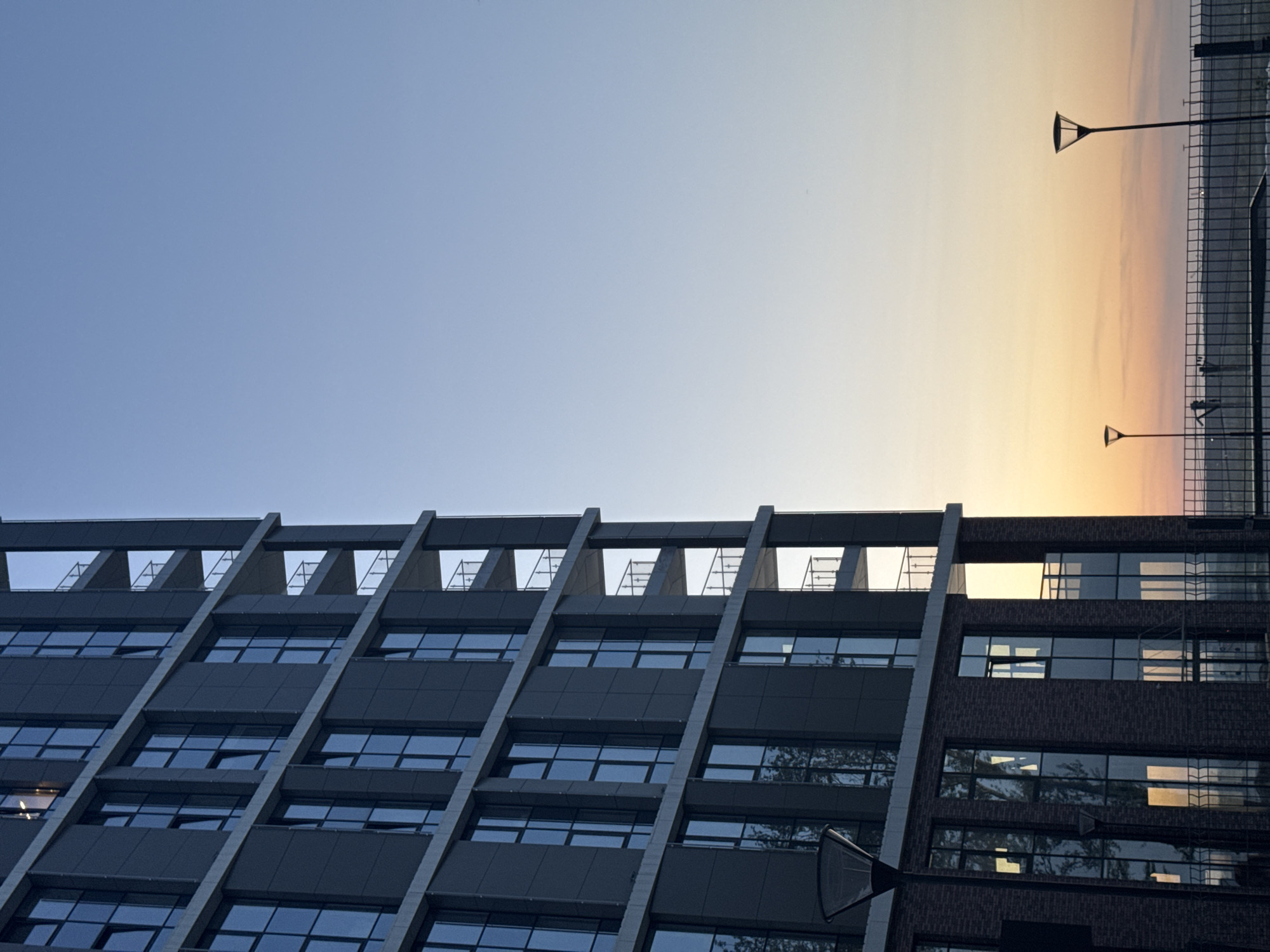The image shows a low-angle view of a modern building in Perm, Russia. The building has a distinctive facade with a grid of concrete beams and rectangular windows. The windows reflect the blue of the sky. Some of the windows are open. The sky transitions from blue to a light orange gradient, suggesting either sunrise or sunset. A fence and two lampposts are visible on the right.
