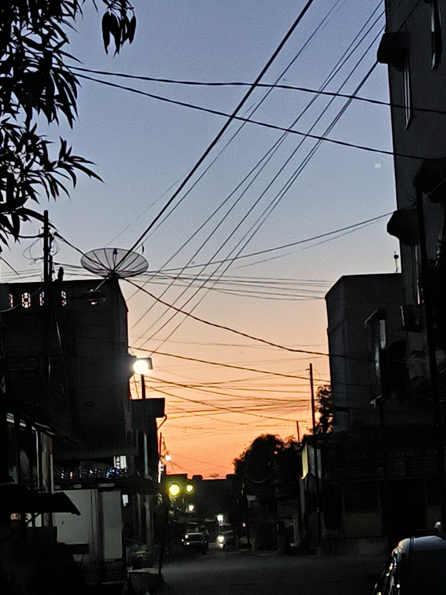 The photograph captures a street in Lhokseumawe, Indonesia, at dusk. The sky transitions from a light blue-gray at the top to a bright orange on the horizon line. Silhouetted buildings flank the street, with a cluster of low-rise structures on either side. Overhead, a tangle of electrical wires crisscrosses the scene. A satellite dish is perched atop one of the buildings on the left. Streetlights illuminate the road, where several cars are traveling. A tree with dense foliage is visible on the left side of the frame. The overall impression is one of a busy, urban environment during the transition from day to night.