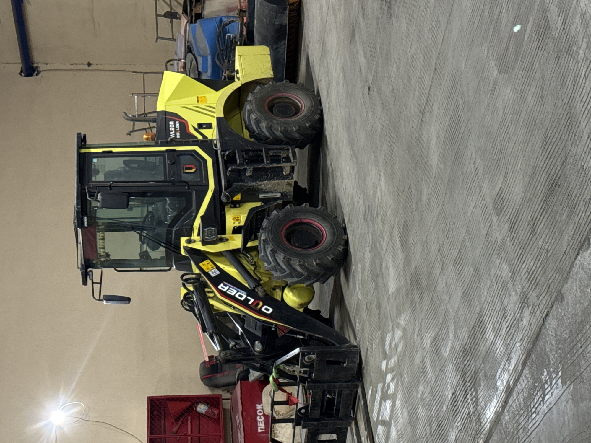 A yellow and black Digger wheel loader is located in an indoor area, possibly a warehouse or storage facility, in Shkotovo, Russia. The loader is partially facing the viewer, showcasing its cab, wheels, and loading arms. In the background, there is additional equipment, including a blue vehicle and red container. The floor is concrete.