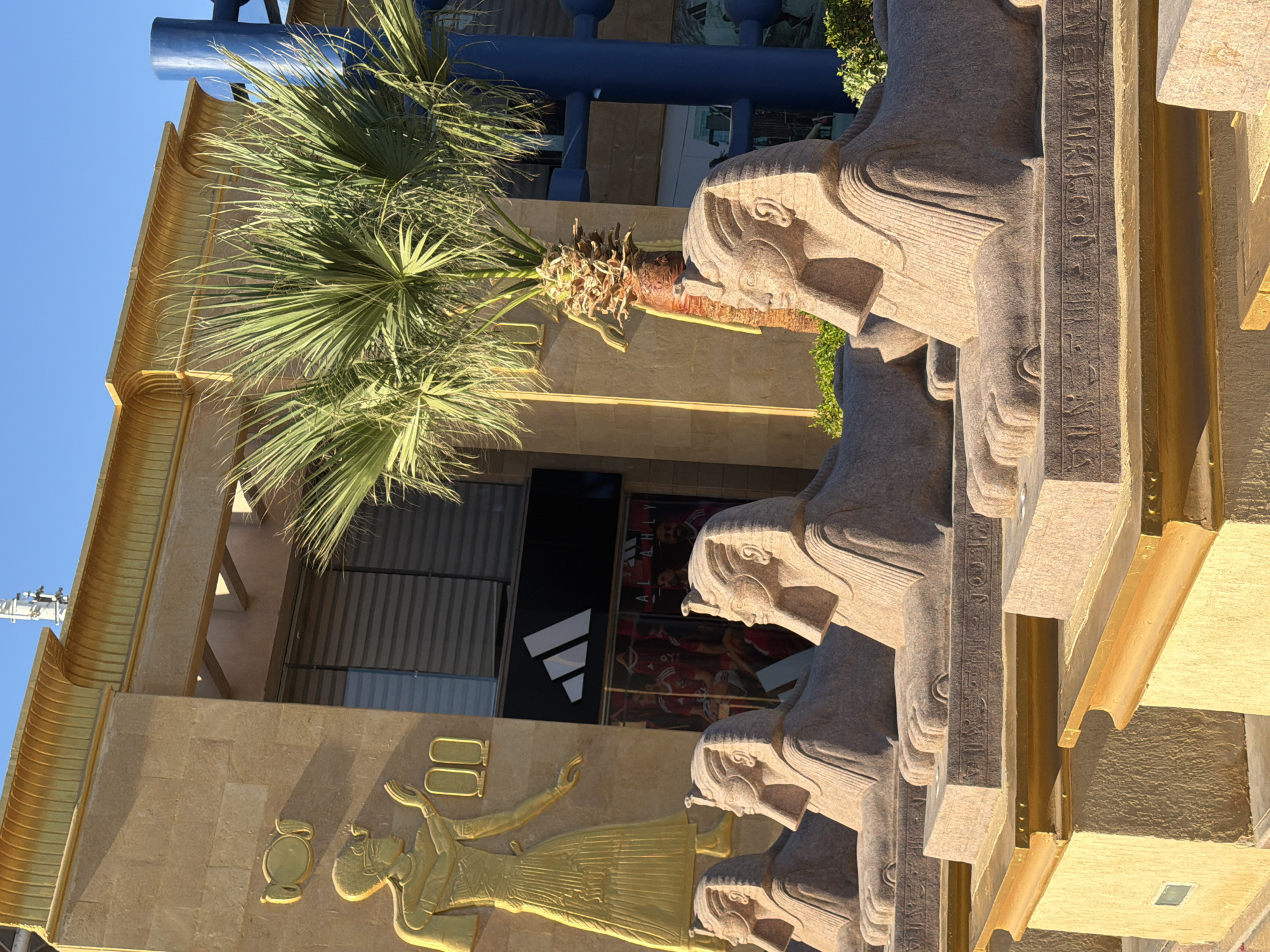 The image shows a row of stone sphinx statues in front of a building in Sharm El Sheikh, Egypt. The building has a facade with Egyptian-inspired motifs, including a golden relief of a pharaoh. There is also an Adidas store visible behind the sphinxes. Palm trees are present in the foreground and background, and the sky is clear and blue. The sphinxes are positioned on raised platforms with hieroglyphic inscriptions. The lighting suggests it is daytime, possibly late afternoon due to the warm tones and shadows. The overall scene evokes a blend of ancient Egyptian aesthetics and modern commercial enterprise.