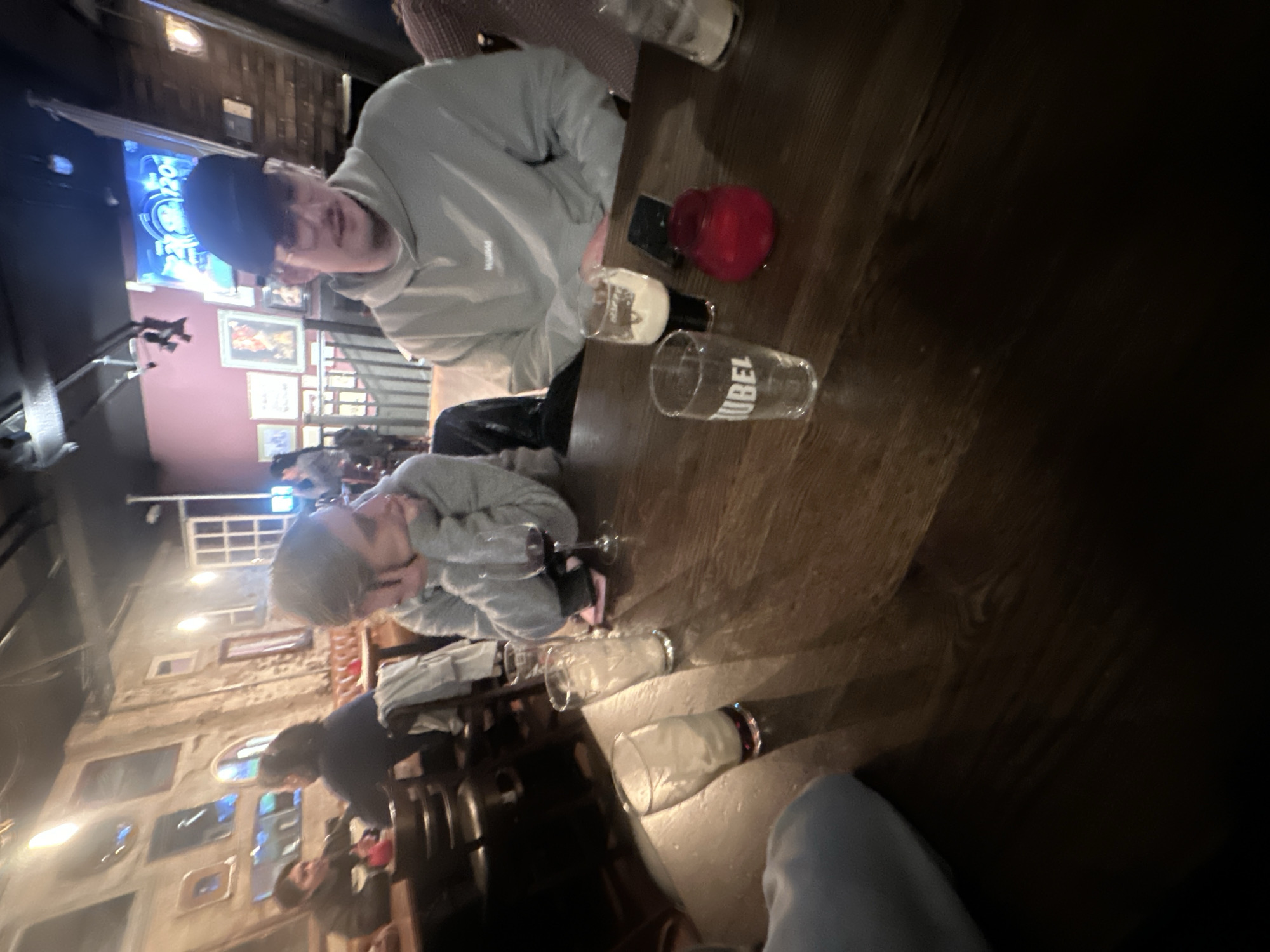 The image shows the interior of a bar or pub, possibly in London, UK. Two people are seated at a dark wooden table, engaged in conversation. On the table are several empty or partially empty beer glasses, a red jar, and a mobile phone. In the background, other patrons are seated at tables and the walls are decorated with multiple framed pictures. A television screen displaying a sports game is also visible in the upper left of the frame. The overall lighting is dim.
