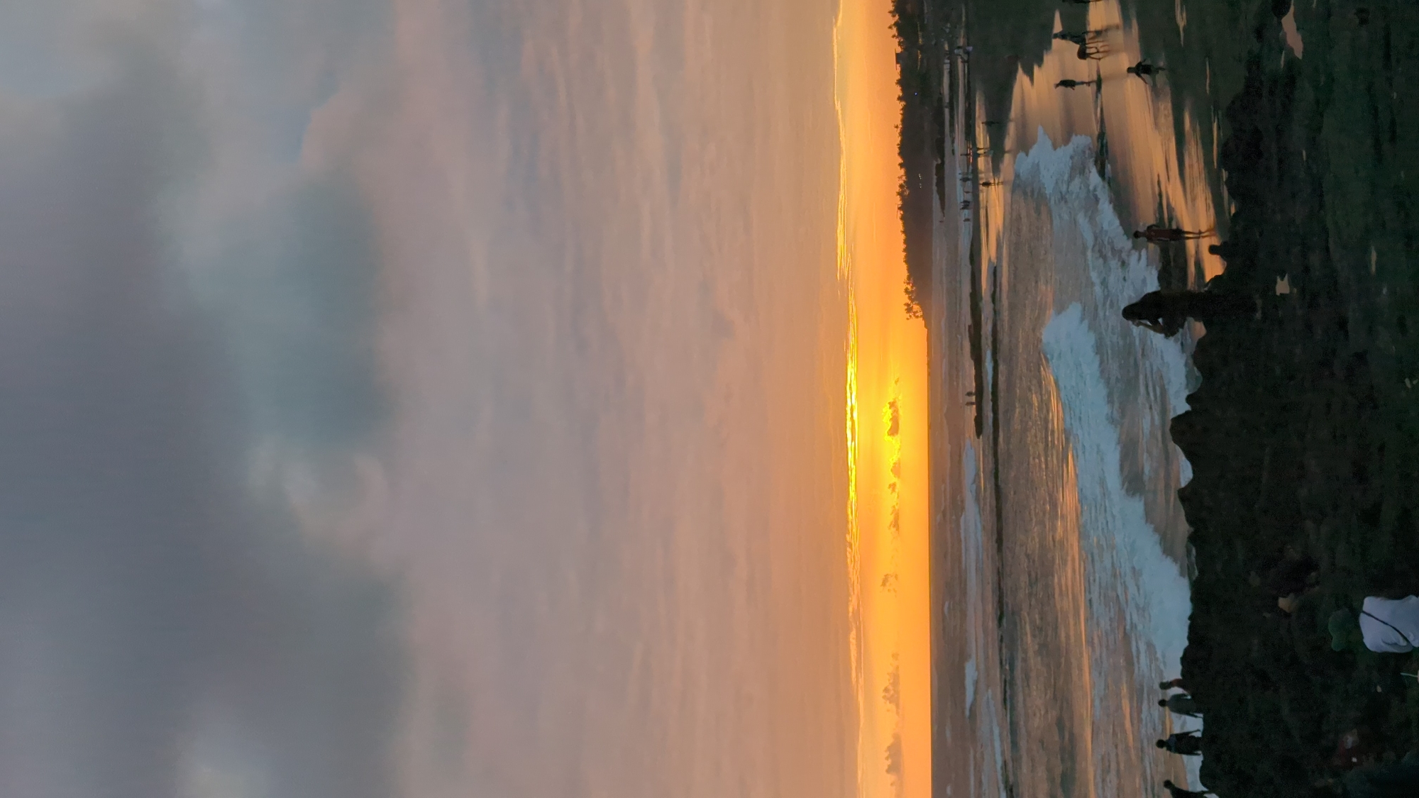 This is a vertical, eye-level photograph of a sunset at Echo Beach in Canggu, Mengwi, Indonesia. The sun is low on the horizon, casting a warm orange glow across the sky and reflecting on the ocean. Dark clouds are present in the upper portion of the sky. Waves are breaking on the shore, and there are several people visible on the beach and in the water. Some individuals are standing on dark, rocky formations in the foreground, while others are walking along the wet sand. Silhouettes of palm trees are visible along the coastline in the distance. The overall scene depicts a serene evening at a popular beach destination.