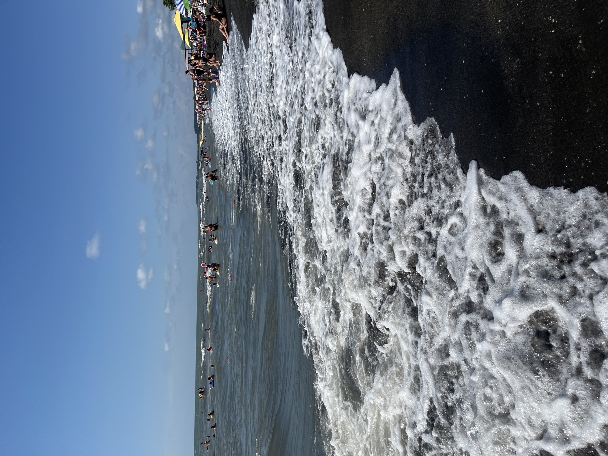 This is a photograph of a crowded beach in Ureki, Georgia, on a sunny day. The foreground is dominated by a wave of white foam washing over dark, fine-grained sand. In the midground, numerous people are visible in the water, some swimming, others wading, and a few using inflatable rings. The water appears choppy with moderate waves. In the background, the beach stretches along the coastline, lined with umbrellas, beach chairs, and more people relaxing or walking. The sky is a clear blue with a few scattered white clouds. The scene depicts a typical summer day at a popular beach resort.