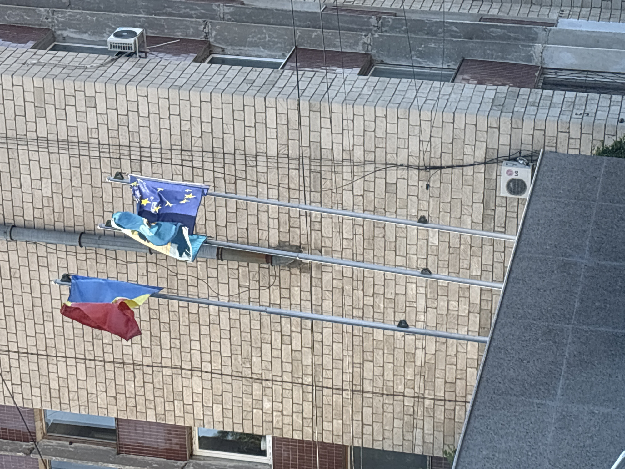 The image shows a brick building with three flags hoisted on poles. The flags appear to be the Romanian flag (red and blue vertical stripes with a yellow emblem), the Ukrainian flag (blue and yellow horizontal stripes), and the European Union flag (blue with yellow stars). An air conditioning unit is visible on the exterior wall of the building. The setting is outdoors, with electrical wires strung across the building. The architecture suggests an urban environment. Given the flags displayed, this location could be a government building or a place with significant symbolic importance in Chișinău, Moldova, potentially related to international relations or European integration efforts.