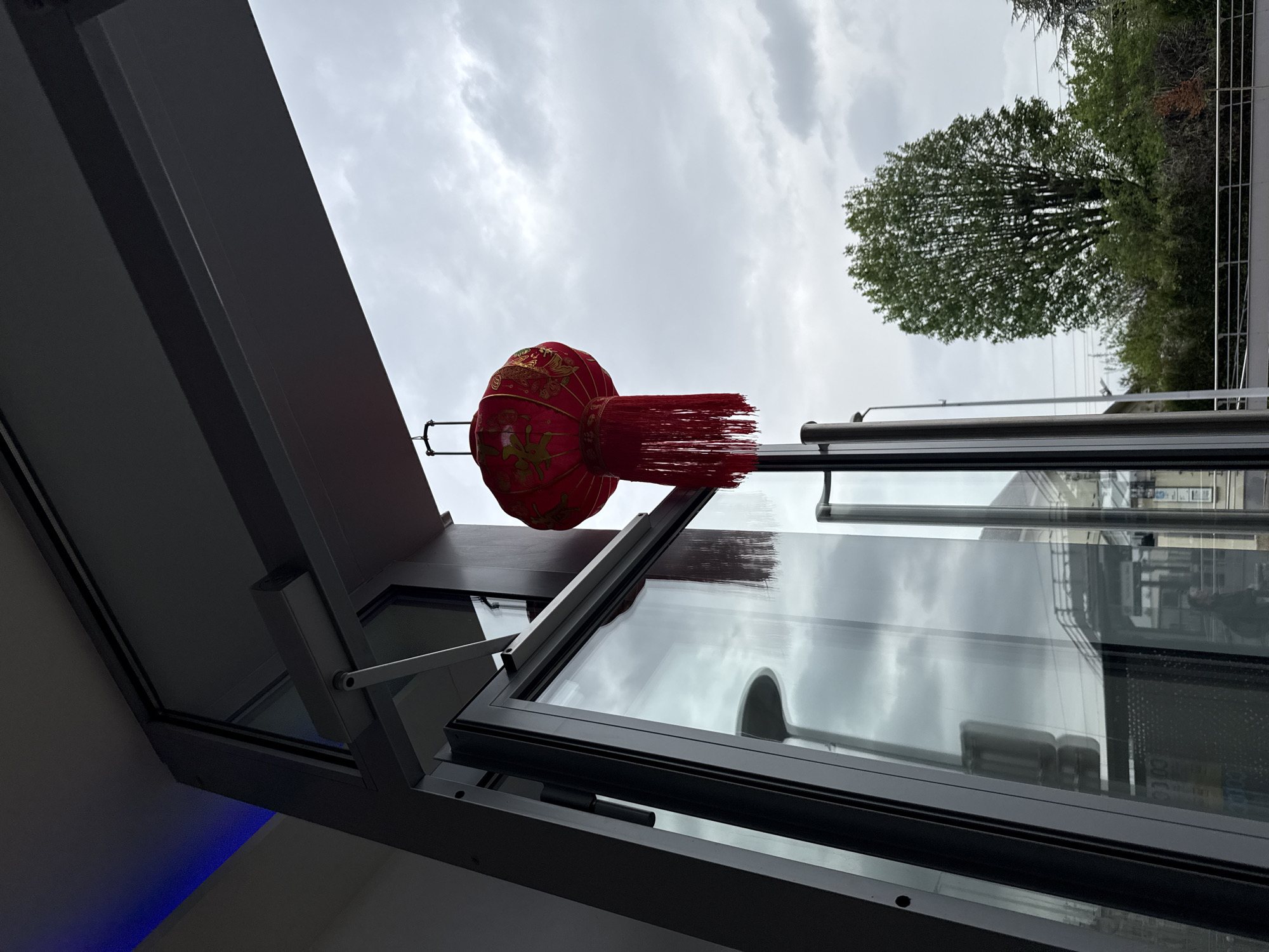 A low-angle, upward shot reveals a red, spherical Chinese lantern with a tassel, suspended by a thin rod from a gray awning. The shot is taken from the city of Domont, France. In the backdrop, the sky is overcast with gray clouds. Behind the lantern, the open window of a building reflects the clouds and surrounding structures, including a tree with green foliage, and the silhouette of a building across the street. The window's frame is dark gray, and the window is slightly ajar. The interior of the building is only vaguely visible, appearing dark. The scene is generally muted in color, except for the vibrant red of the lantern.