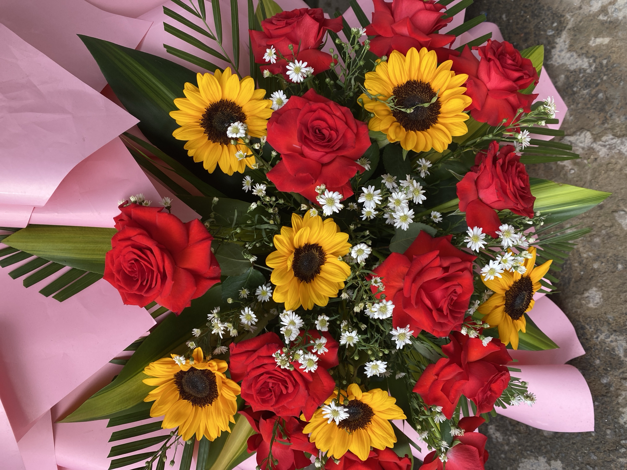 A close-up, overhead shot shows a large floral arrangement composed of red roses, sunflowers, and small white filler flowers, all set against a backdrop of pink paper wrapping and green foliage. The bouquet is placed on a rough, gray surface that appears to be concrete. The image is well-lit and the colors are vibrant. The scene does not depict any people, actions, or interactions. The location is not identifiable as being in the city of Da Nang, Vietnam, or any other specific location.