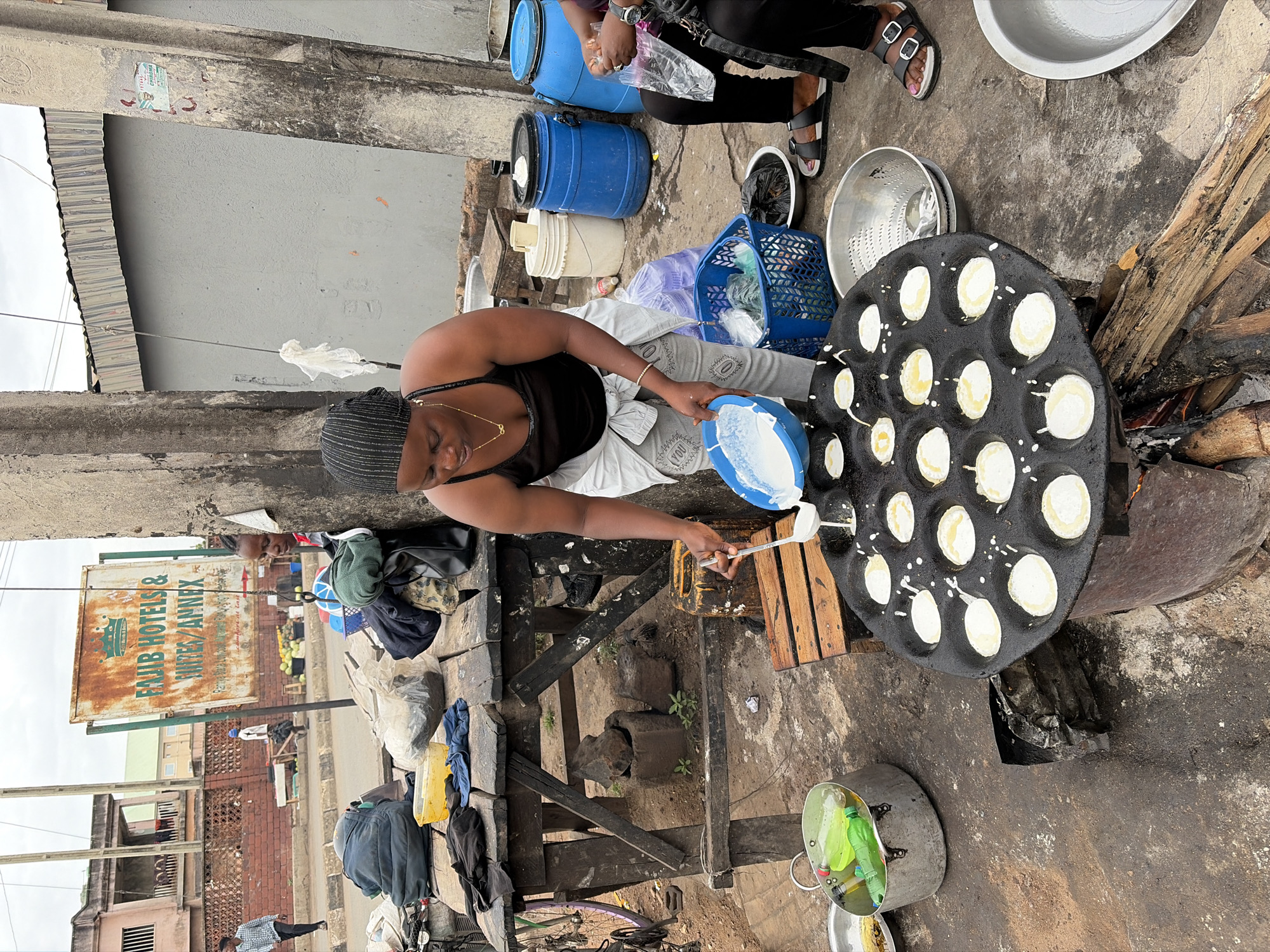 An outdoor scene in Jos, Nigeria, shows a woman wearing a black tank top, grey pants, and a striped head covering while cooking. She is pouring a white batter from a blue bowl into a round, black pan with multiple circular indentations, each filled with the batter. The pan is positioned over a fire built within a metal container, with firewood scattered nearby. Other items include blue barrels, plastic containers, and metal bowls surrounding the cooking area. In the background, buildings and a weathered sign for "FAJIB HOTELS & INNER ANNEX" are visible along a street, suggesting an urban setting.